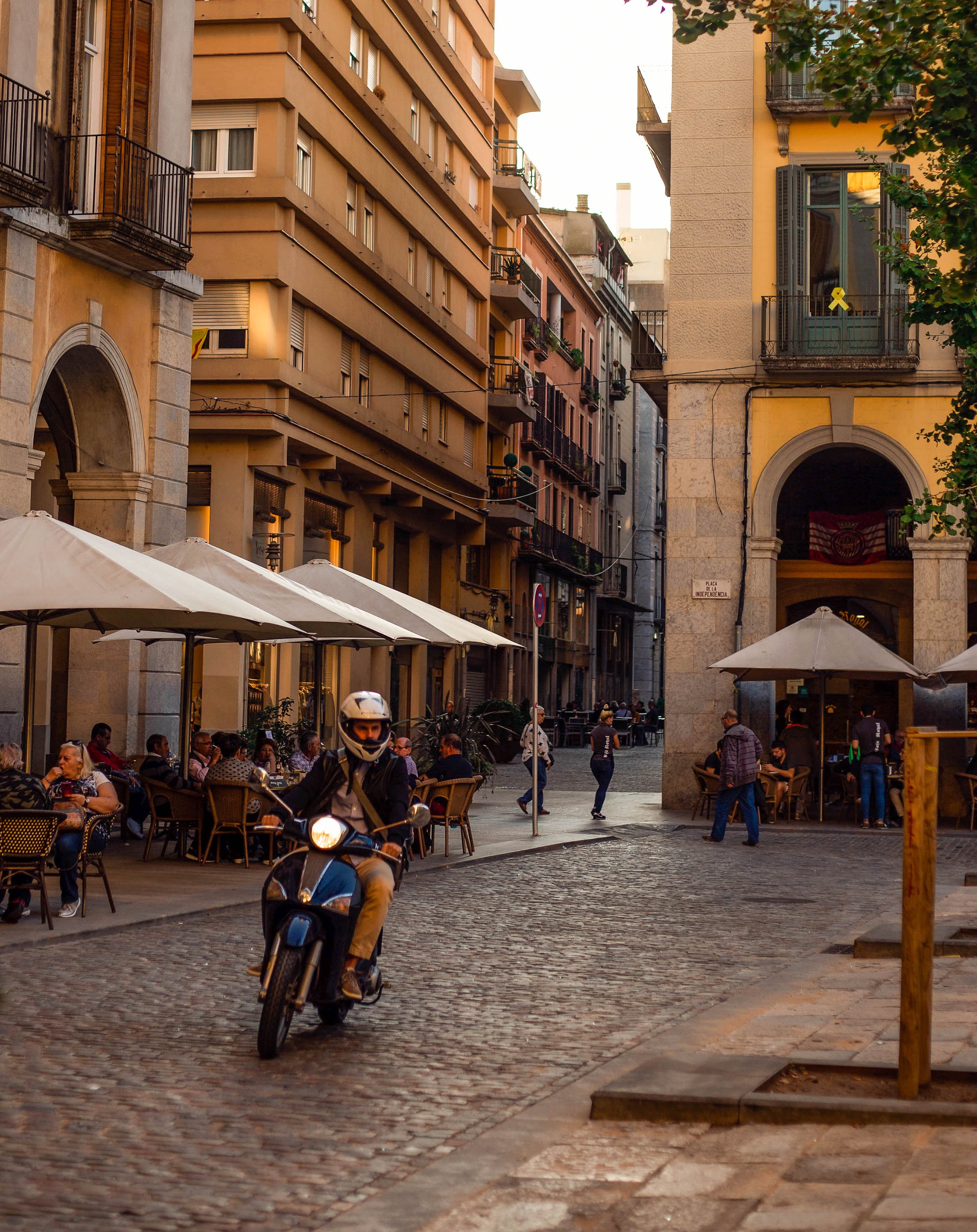 A lively city street scene during the evening with people sitting at outdoor café tables, a person riding a motorcycle, and tall buildings lining the street. The buildings have balconies and are illuminated by warm lighting.
