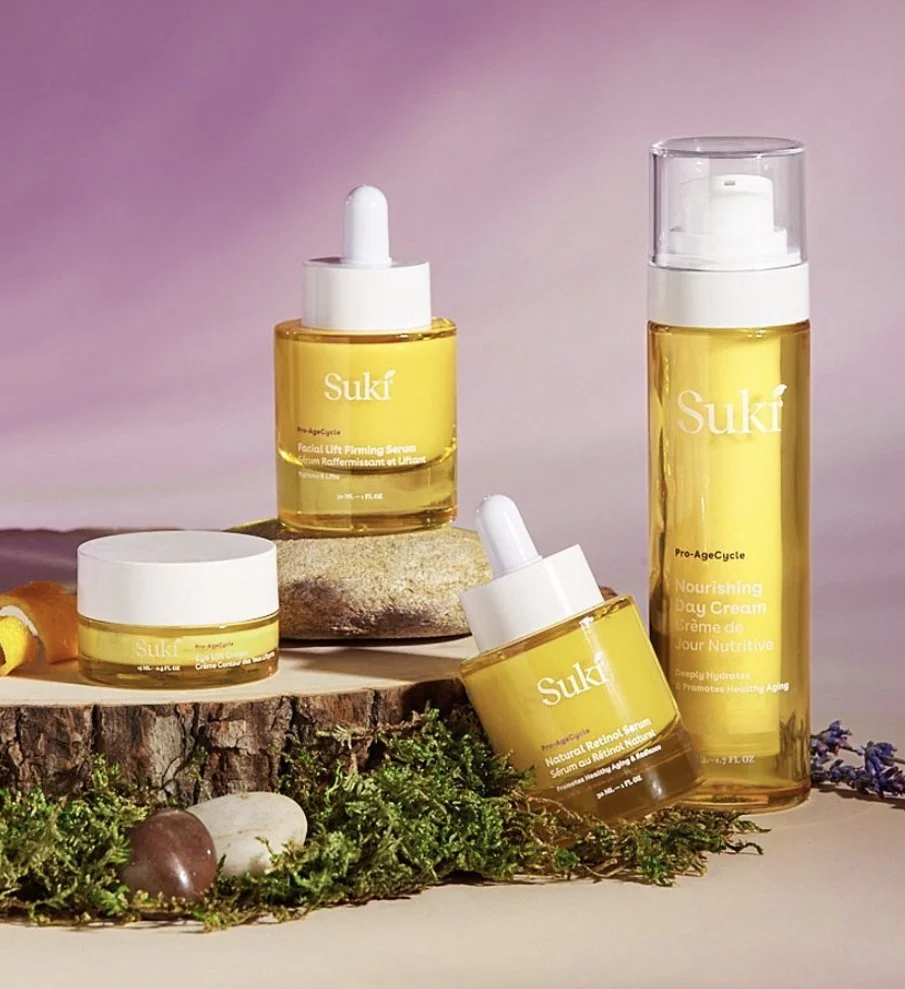 Four bottles of Suki skincare products with yellow liquid in various sizes, arranged on and around a tree trunk slice and natural stones, with green plants and purple flowers in the foreground, against a pink background.
