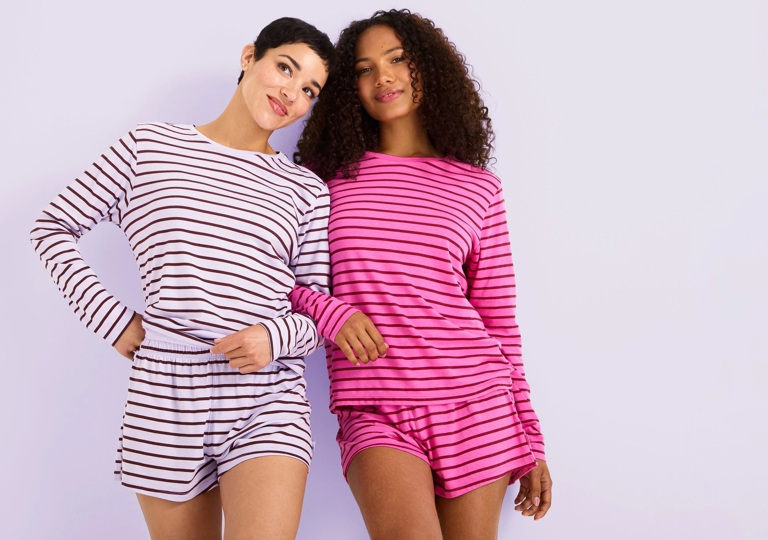 Two women standing side by side, wearing striped loungewear sets, one in white with black stripes and the other in pink with black stripes, smiling softly against a light purple background.