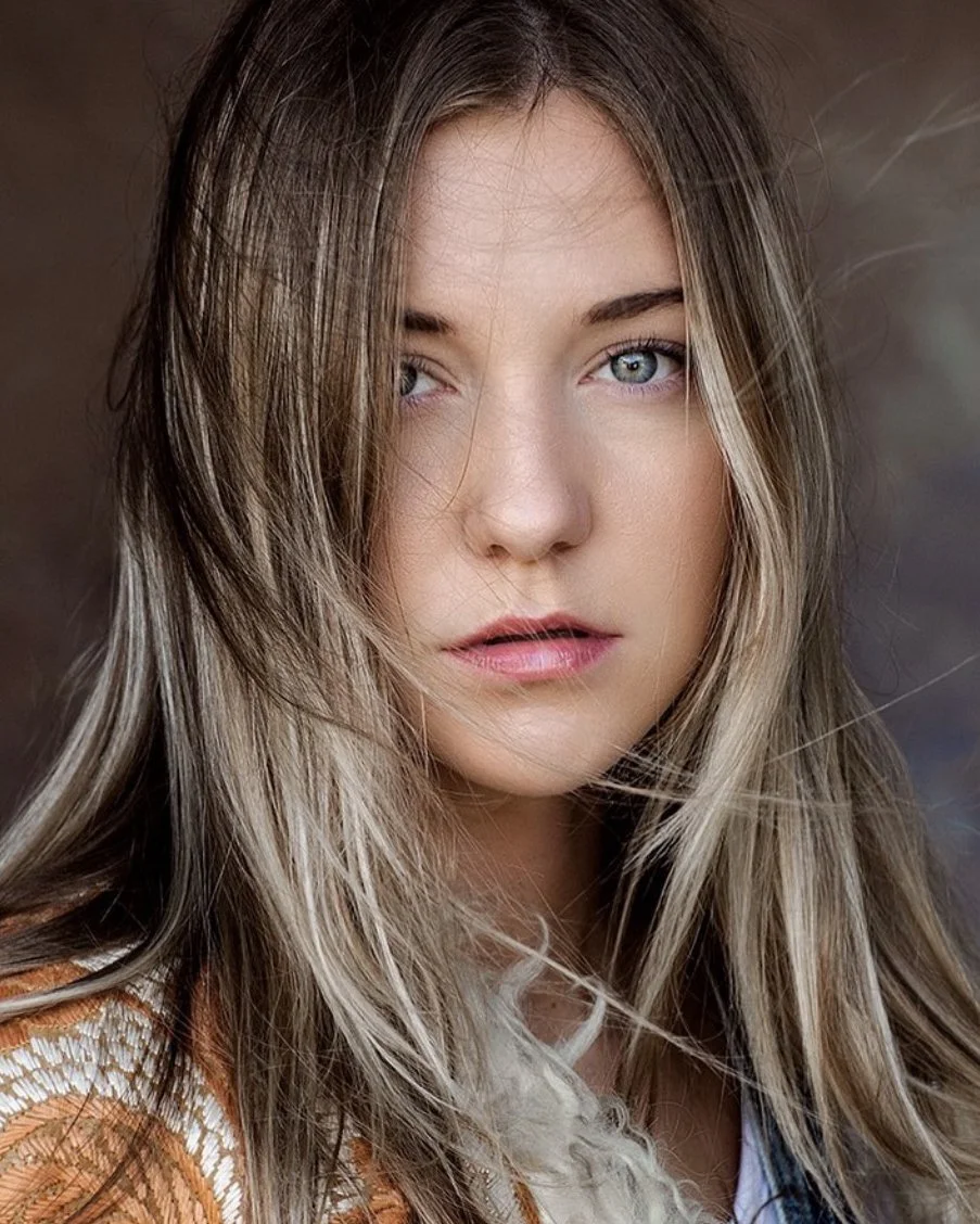 Close-up of a young woman with long, wavy blonde hair and blue eyes, looking slightly to the side with a serious expression, with a blurred neutral background.