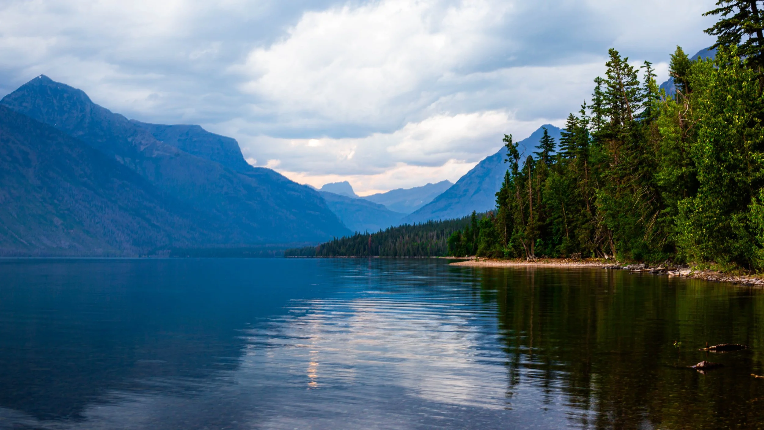 A scenic view of a lake with calm water, surrounded by dense green trees on the right and a mountain range in the background under a cloudy sky.