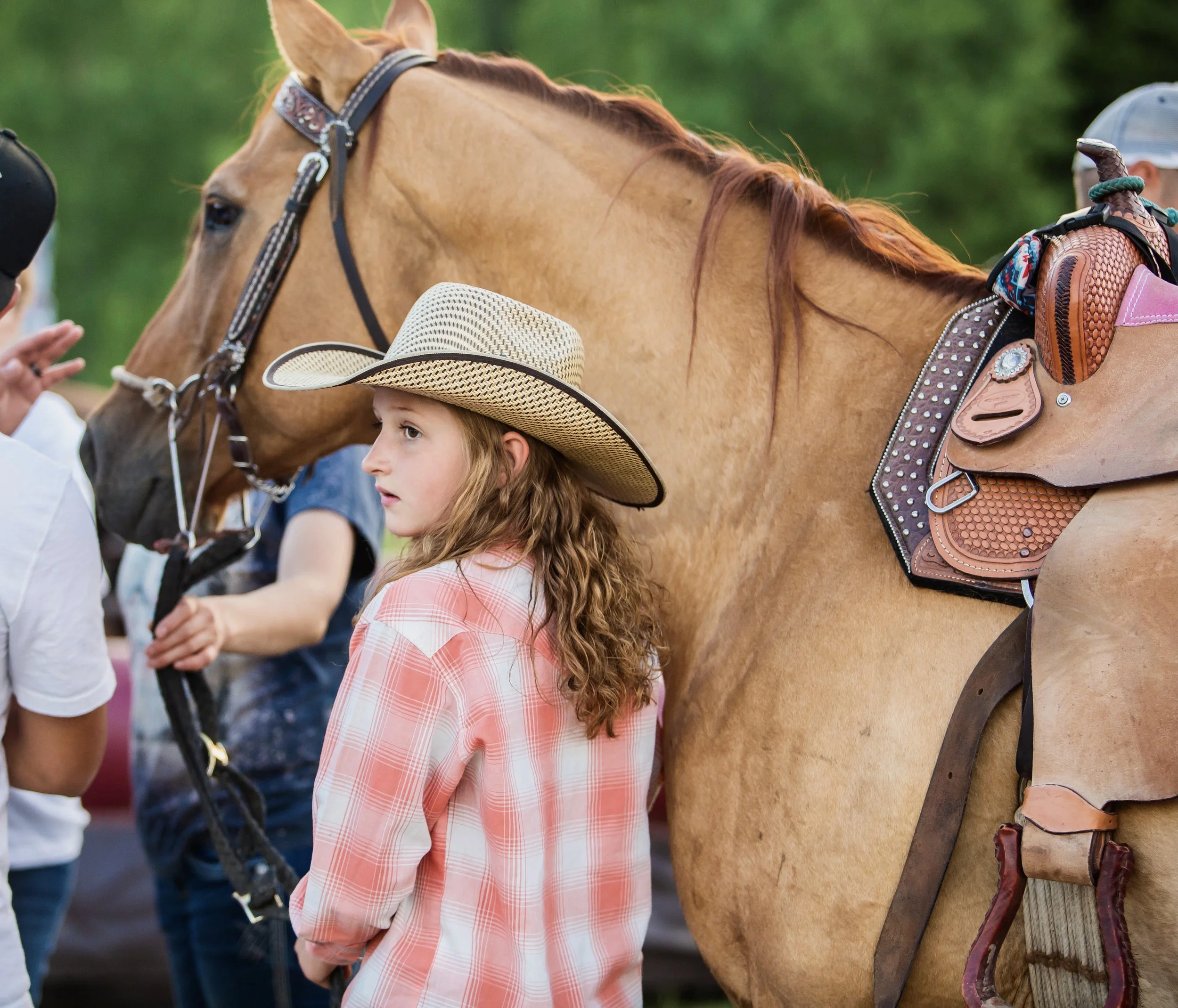 A young girl with long red hair wearing a plaid shirt and a straw hat, standing next to a brown horse with saddlebags, in an outdoor setting with green foliage.