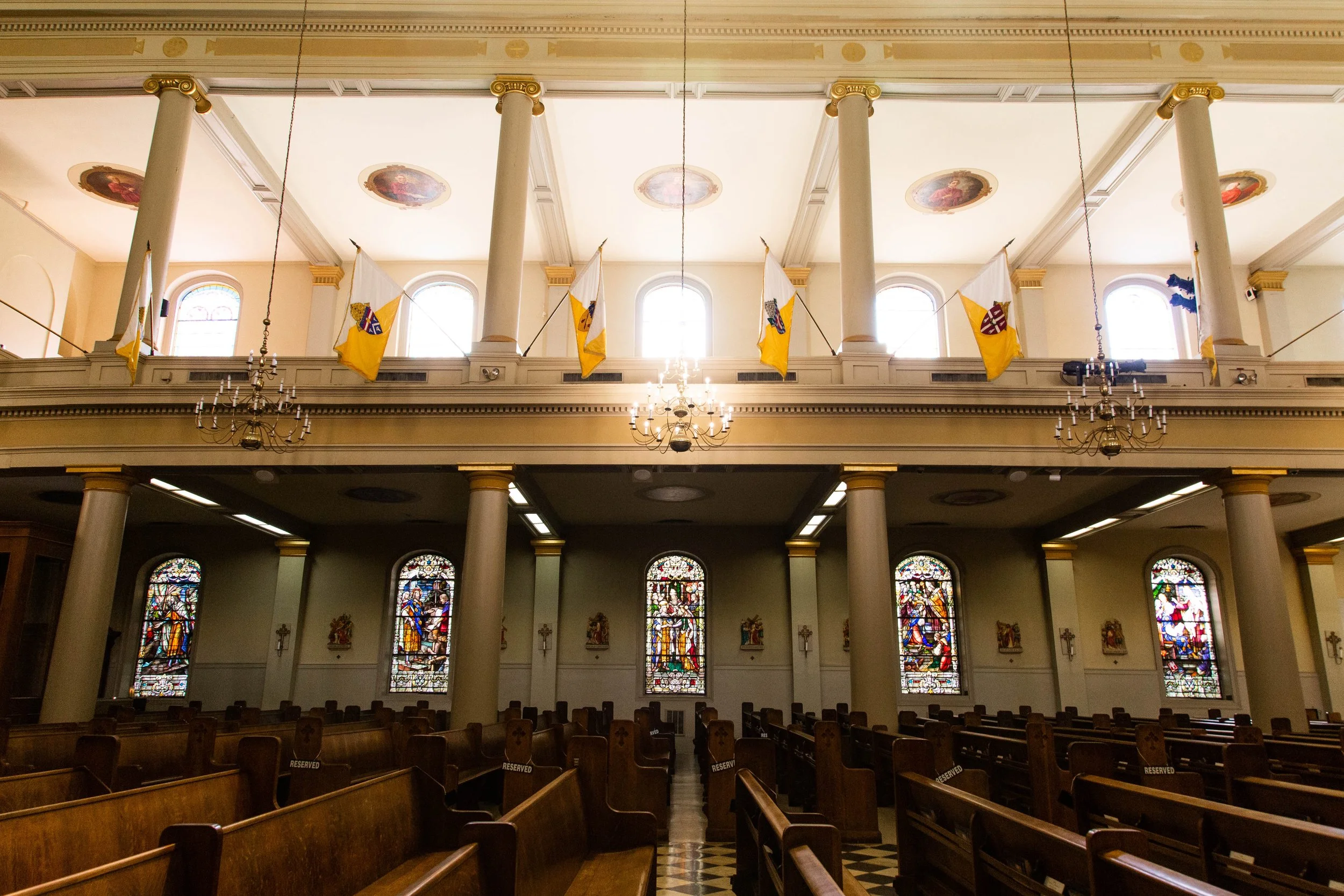 Interior of a church with high ceiling, stained glass windows, chandeliers, white columns, and flags hanging from the upper balcony.