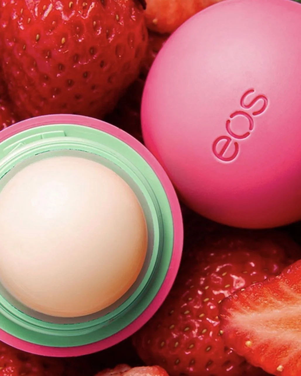 Close-up of a pink EOS lip balm container, with the lid removed revealing the balm inside, surrounded by fresh strawberries.