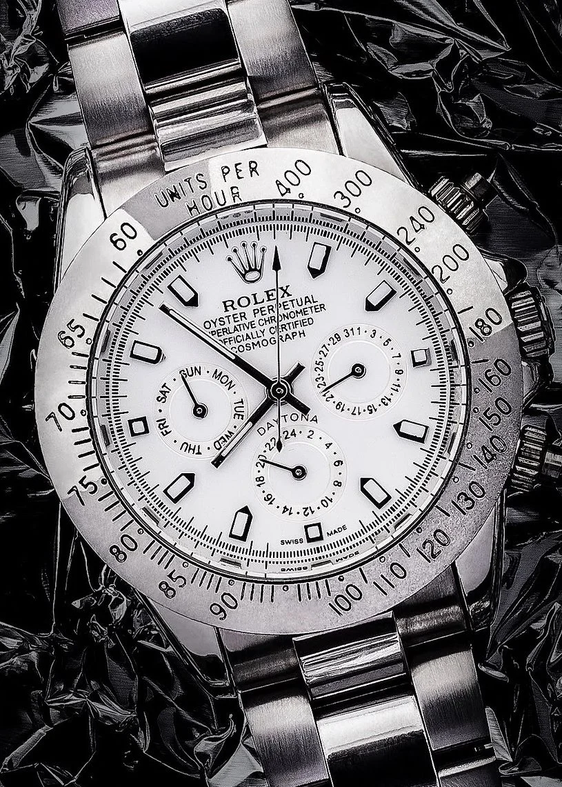 A close-up photo of a stainless steel Rolex Oyster Perpetual Cosmograph watch with a white dial, black hands, and multiple subdials, placed on crinkled black plastic.