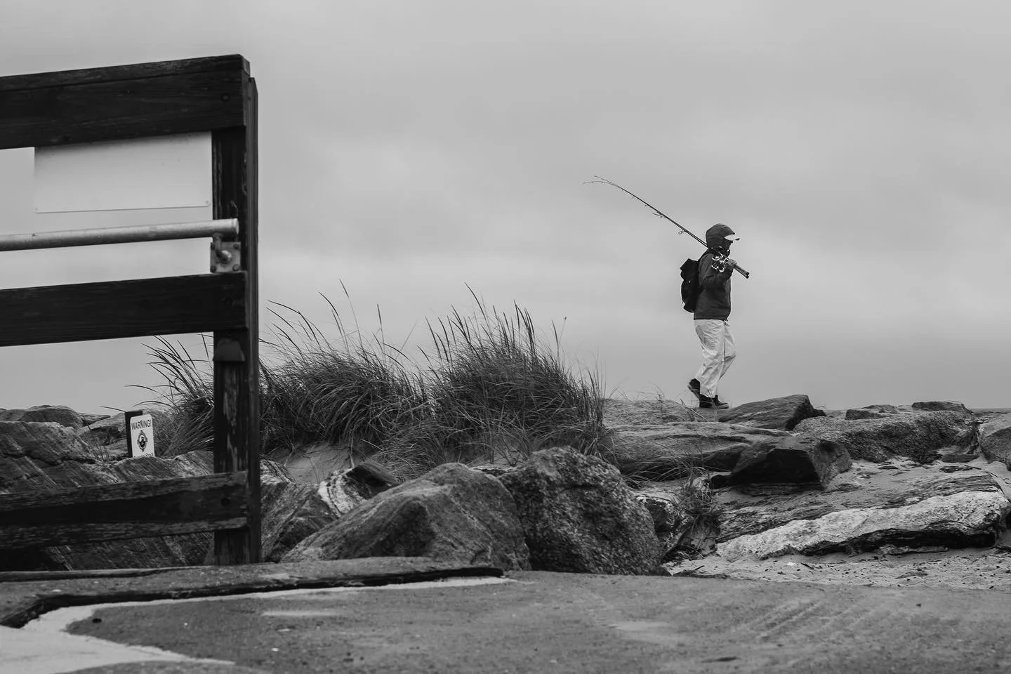 A person in a jacket, pants, and helmet, carrying a backpack, walks on rocks near a beach with a fishing rod over their shoulder. The scene is cloudy, with dunes and grass in the background, and a partially visible wooden structure on the left.
