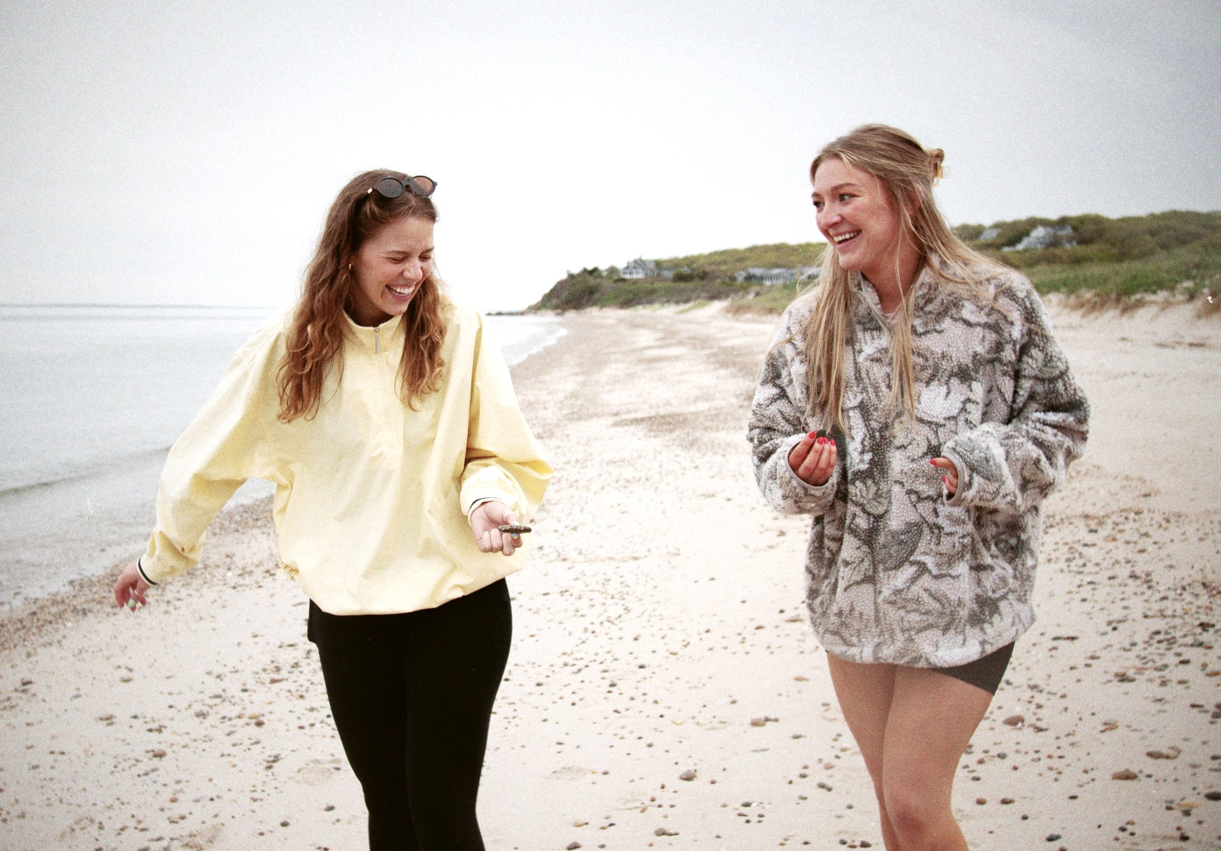 Two women are walking and smiling along a beach with houses on the hillside in the background. One woman has long curly hair and is wearing a yellow jacket and black pants, holding a small object in her hand. The other woman has long straight hair an