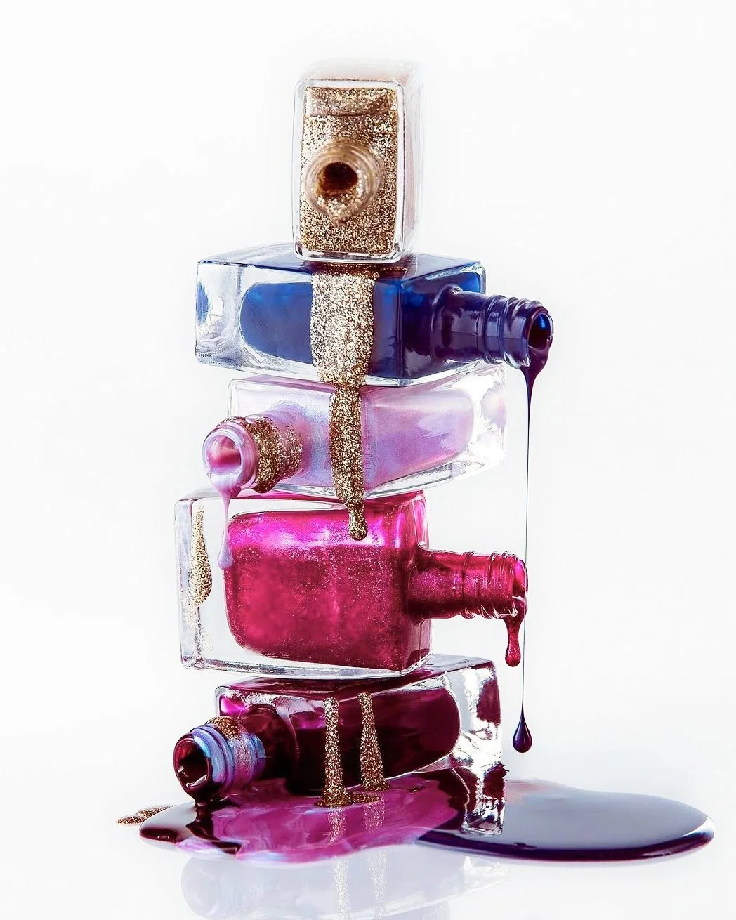 Stacked open nail polish bottles with spilled pink, purple, and gold glitter nail polish leaking and flowing on a white background.