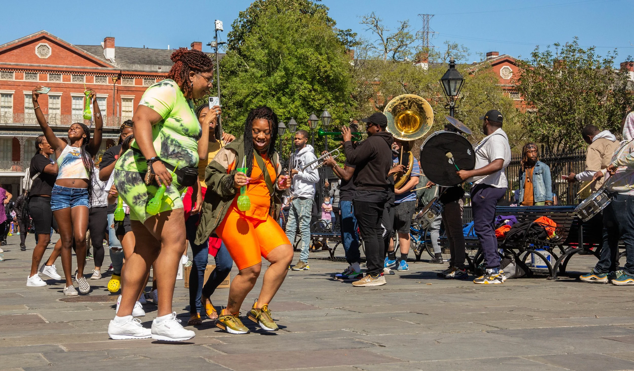 People dancing and listening to a live band playing musical instruments in an outdoor urban setting with trees and red brick buildings in the background.