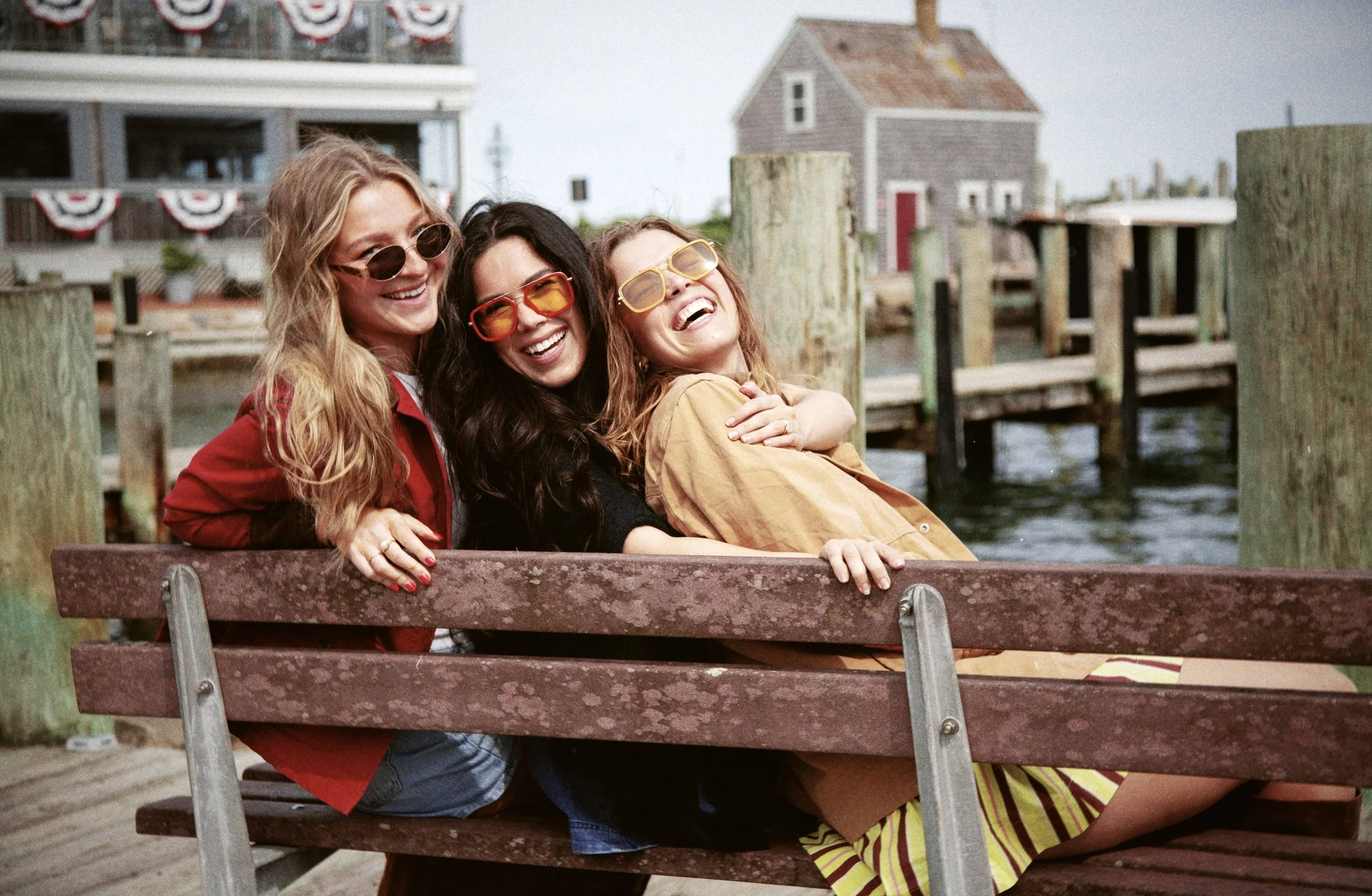 Three women wearing sunglasses sitting on a wooden bench at a waterfront location, smiling and laughing with a small house and a building decorated with red, white, and blue bunting in the background.