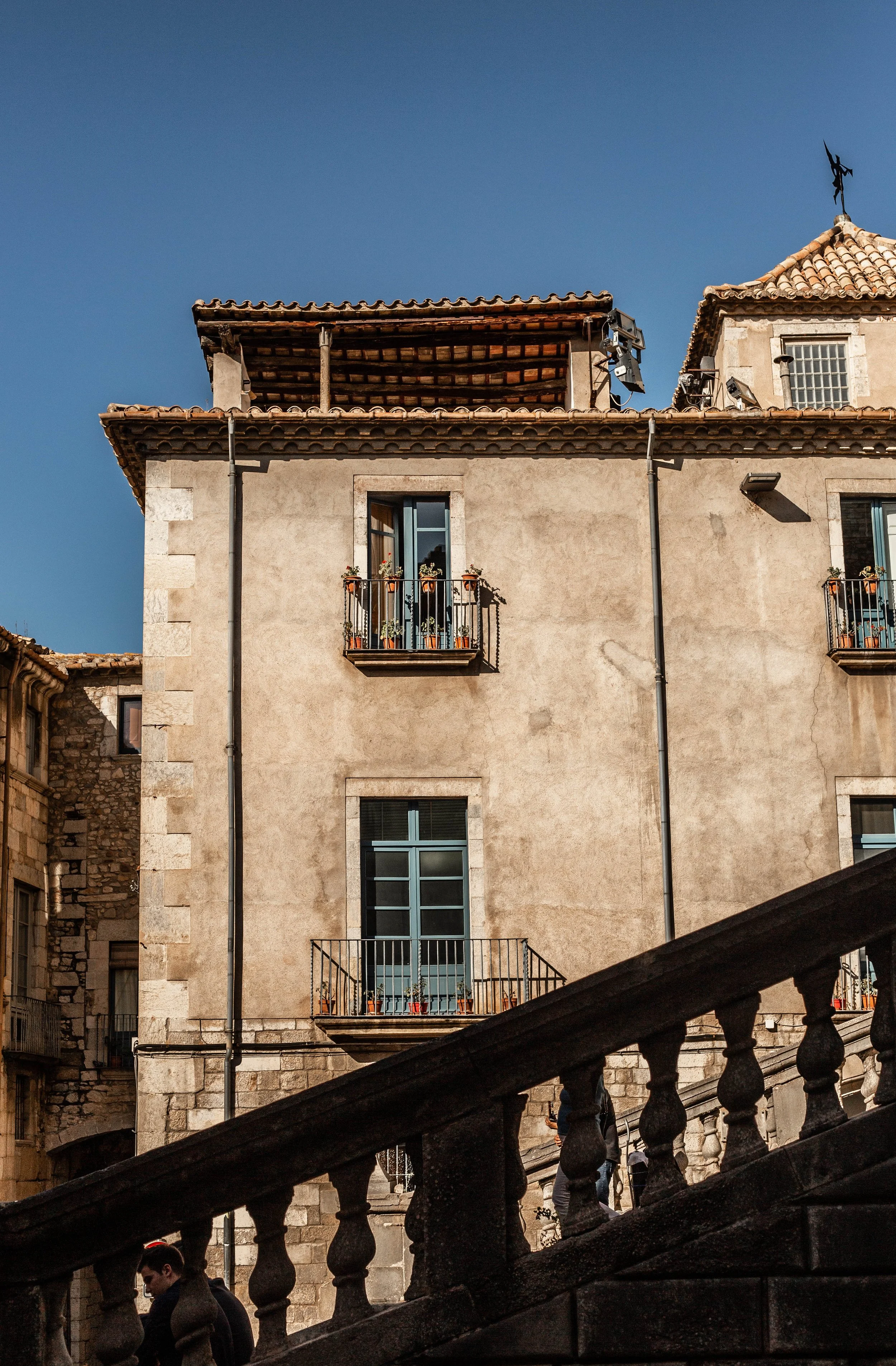 An old European building with balconies, flowerpots, and a terracotta tiled roof, viewed from a stairway with a balustrade in the foreground.