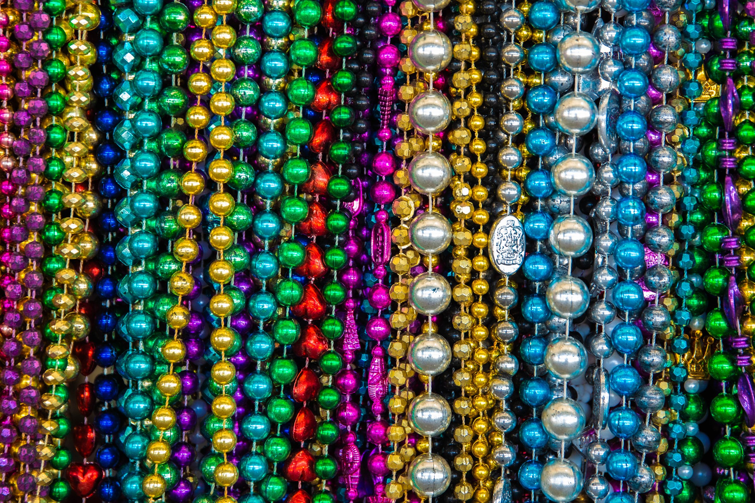 A colorful assortment of shiny Mardi Gras beads including purple, green, gold, red, blue, silver, and black beads arranged in rows.