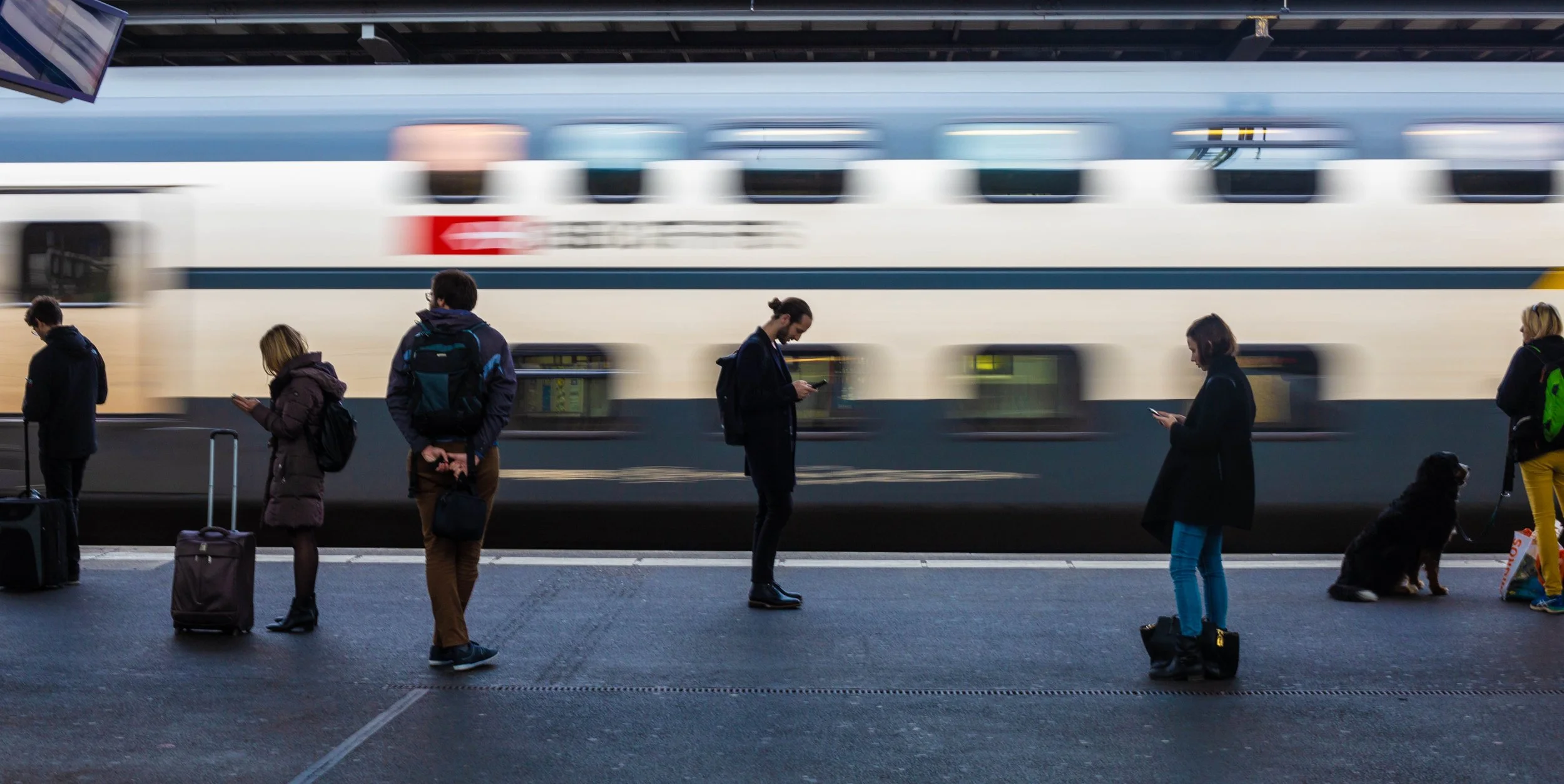 People standing on a train platform waiting for a train, with a train passing by in motion in the background.
