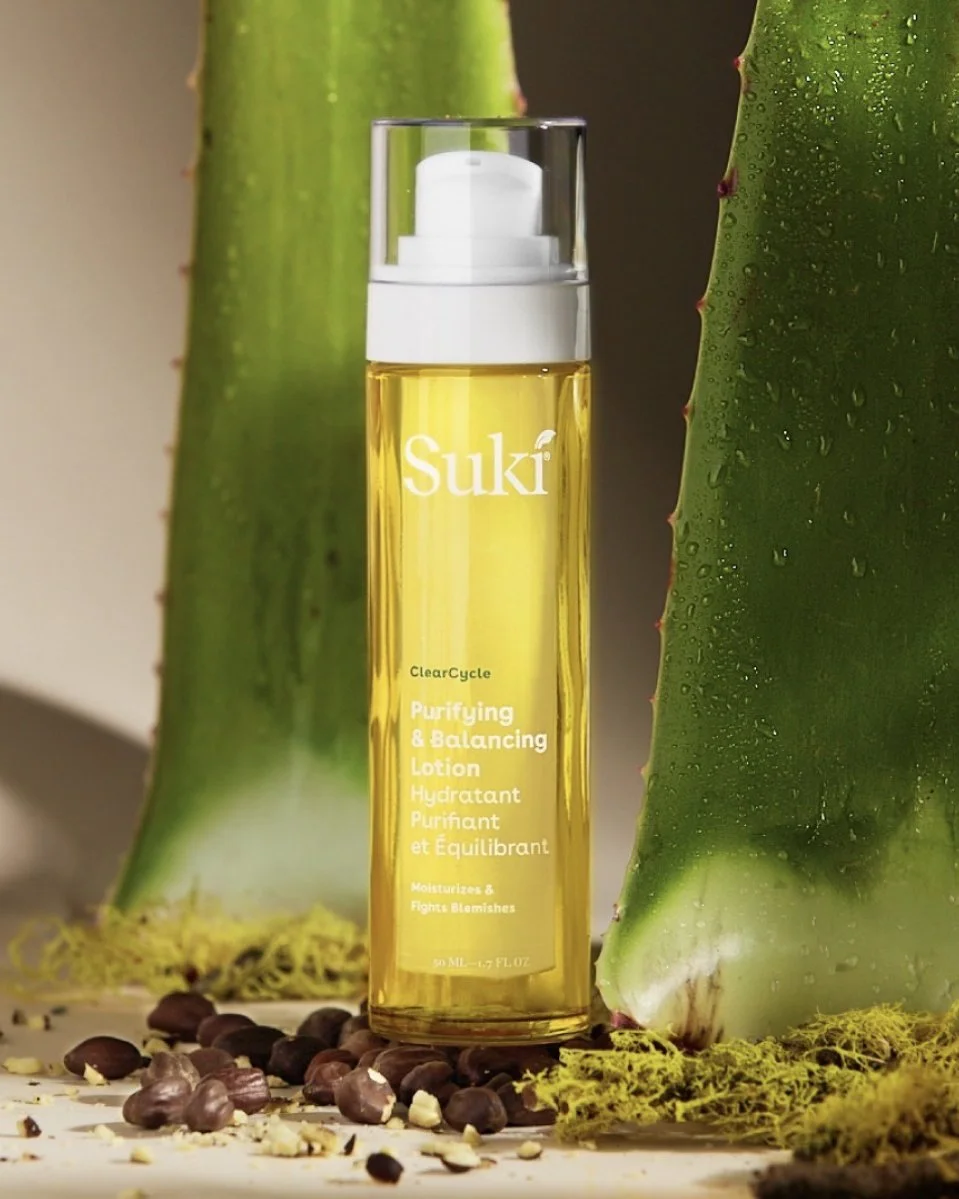 A bottle of Suki Purifying & Balancing Lotion is placed between two large aloe vera leaves with water droplets. The scene includes scattered seeds and moss on the surface.