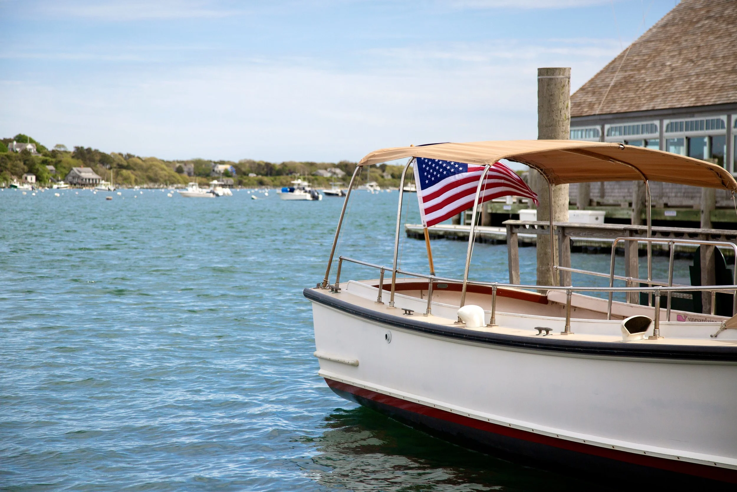 A white boat docked at a wooden pier with an American flag flying on it, in a harbor with multiple boats and houses along the shoreline under a blue sky.