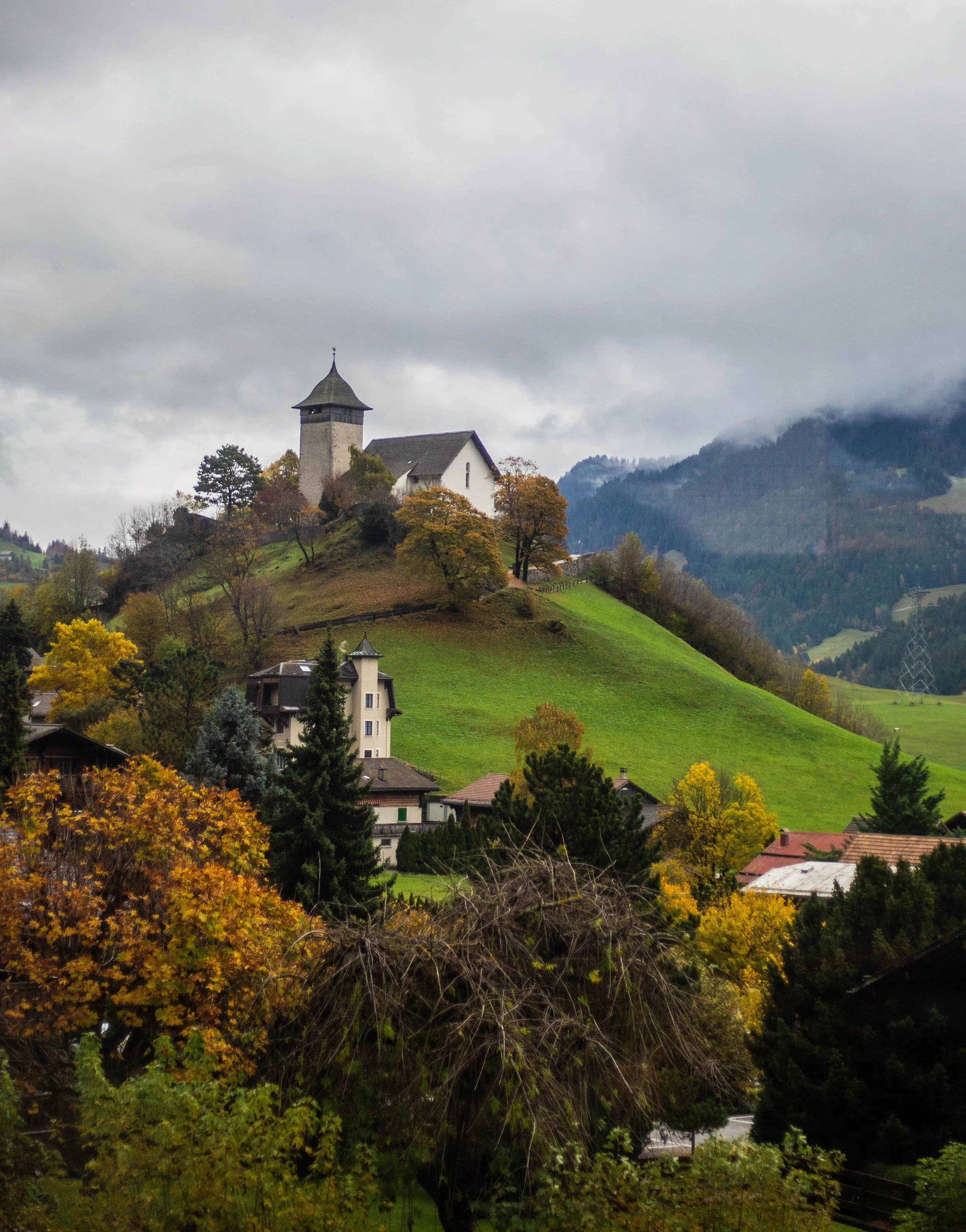 A small church with a pointed steeple located on a hilltop, surrounded by trees with autumn foliage, overlooking a valley with houses and green fields, under a cloudy sky.