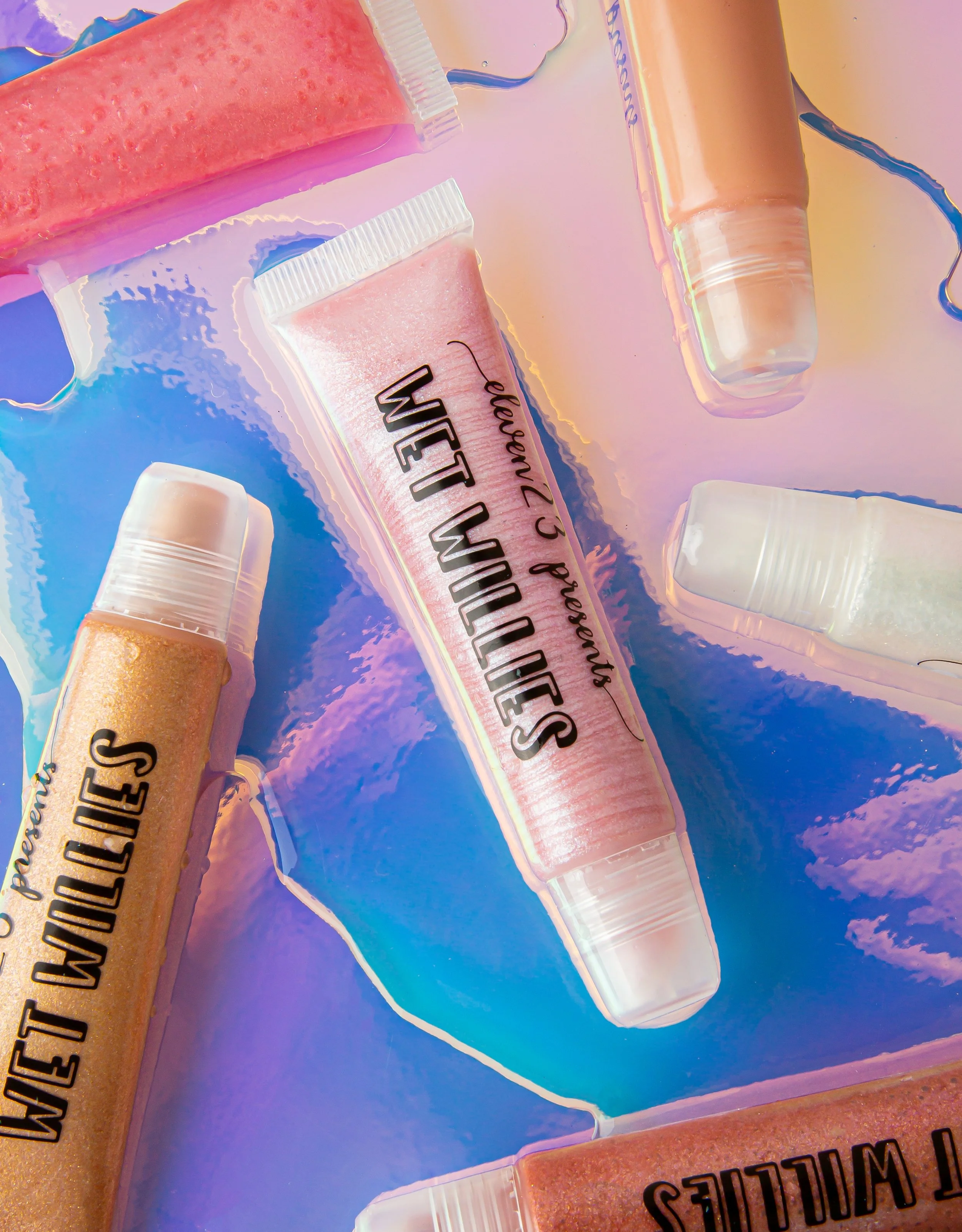 Several makeup or cosmetic products, including lip gloss or lipstick tubes and a container of shimmery powder, arranged on a pastel-colored, iridescent surface.