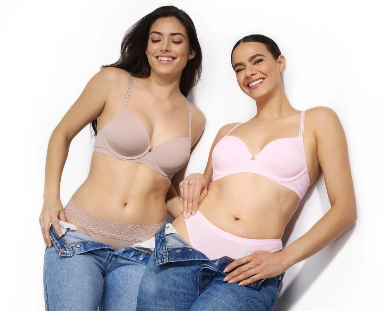 Two women smiling and holding their bras and underwear, wearing jeans against a plain white background.