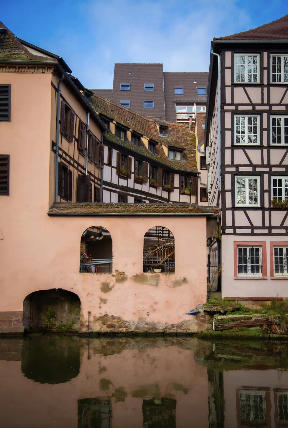 Colorful European buildings with traditional wooden frames, window shutters, and tiled roofs, reflected in a calm river in the foreground.
