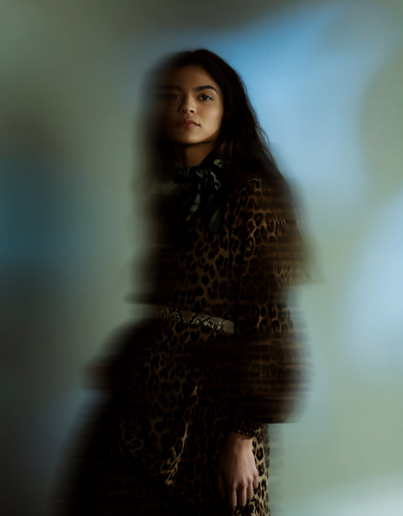 A woman with long dark hair wearing a leopard print outfit, standing against a blurred green and blue background, with a soft focus effect.