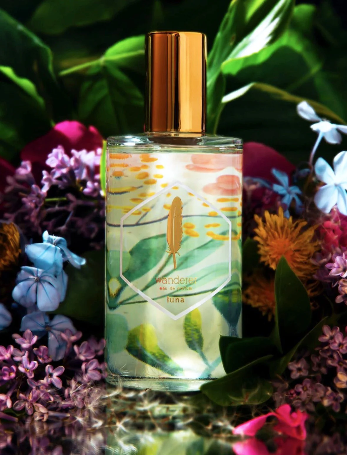 A perfume bottle labeled 'Wanderer Luna' with a gold cap, surrounded by colorful flowers and green leaves.