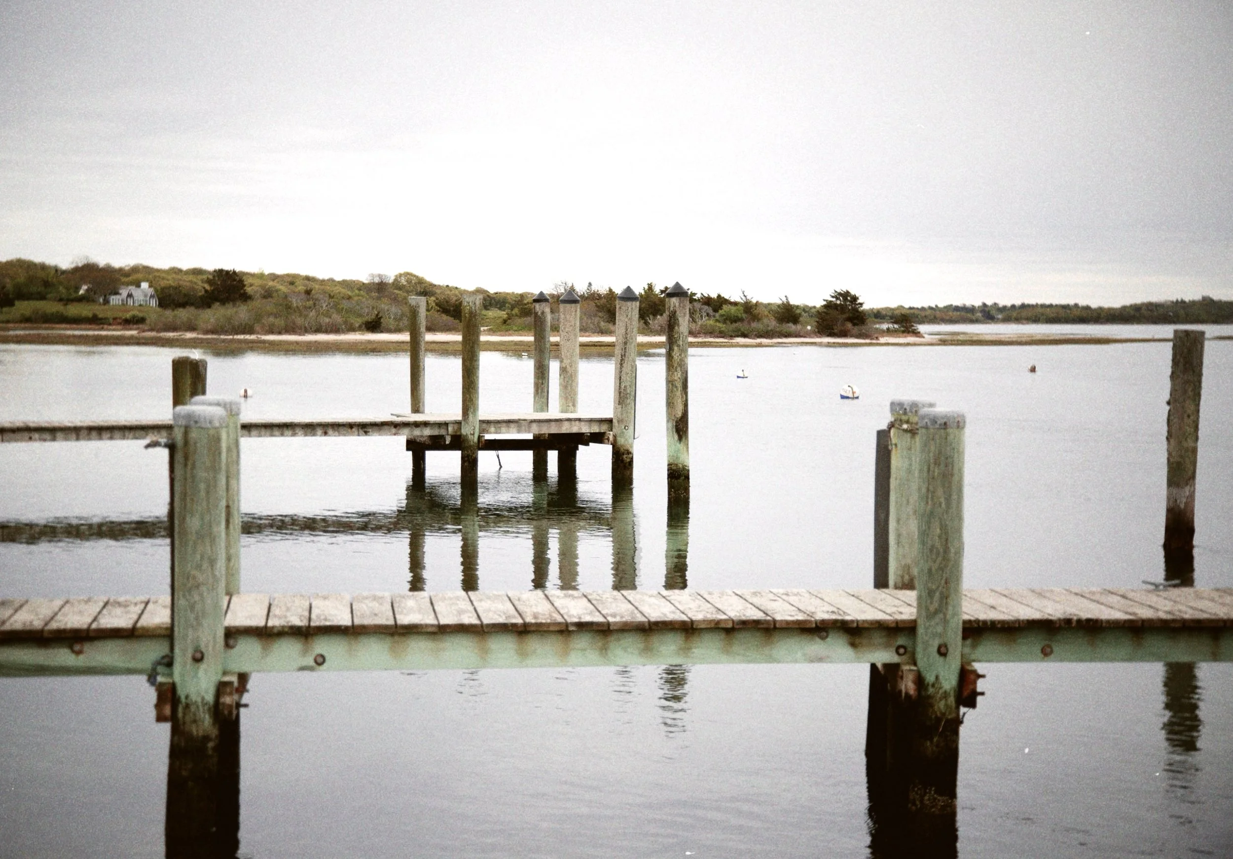 View of a wooden dock extending over the water with a shoreline and trees in the background, under an overcast sky.