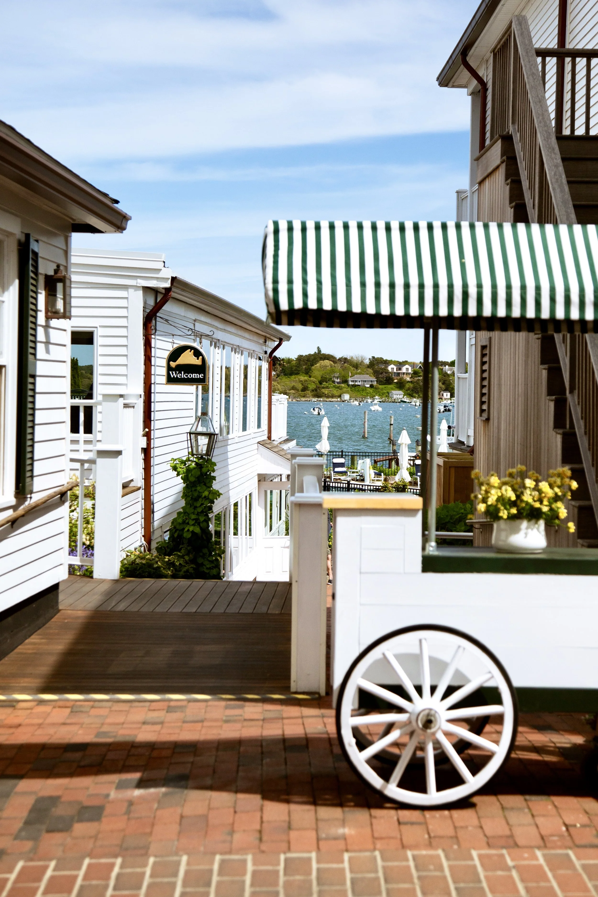 A white wooden cart with a green and white striped canopy, white wheels, and potted yellow flowers, set on a brick patio near waterfront houses and boats.