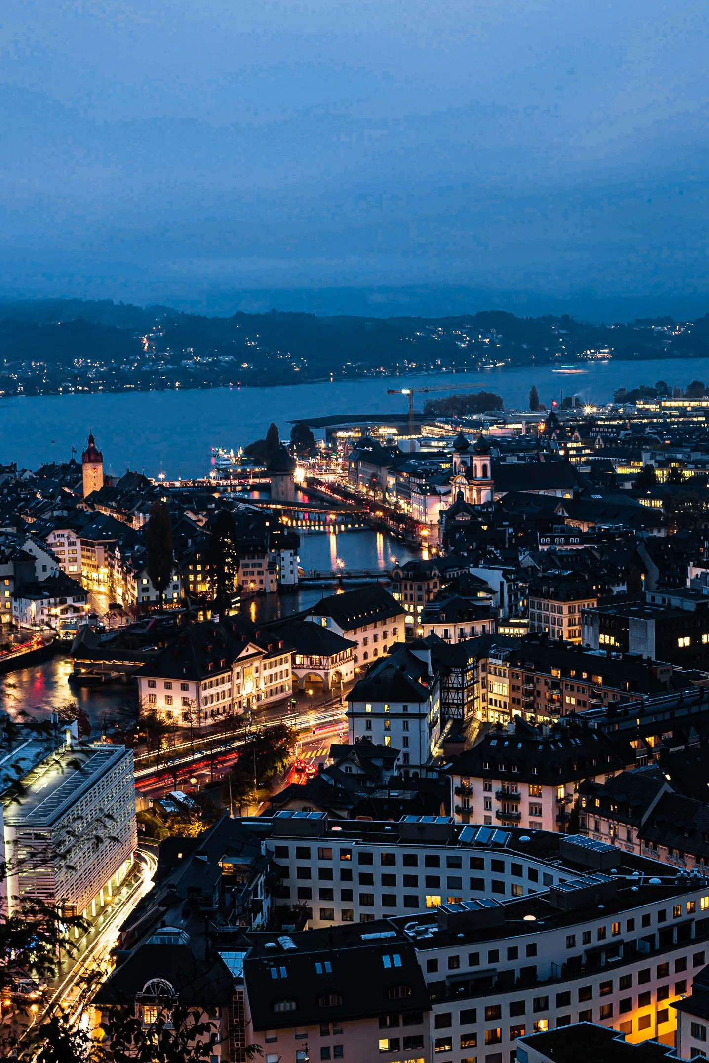 Nighttime cityscape featuring a river, illuminated buildings, and distant hills under a dark blue sky.