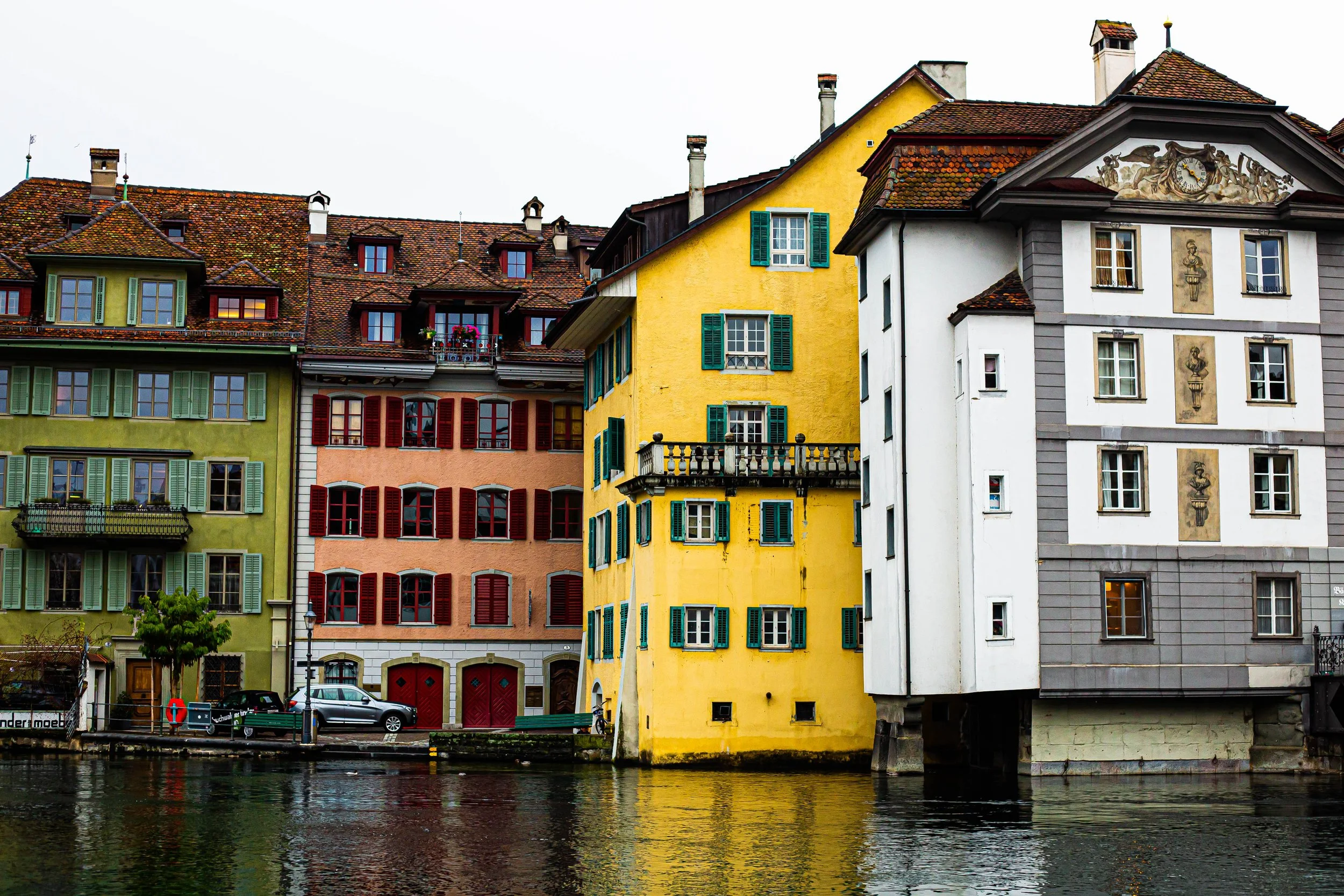 Colorful European-style buildings alongside a river with reflections, featuring green, peach, yellow, and gray facades, red shutters, and tiled roofs.