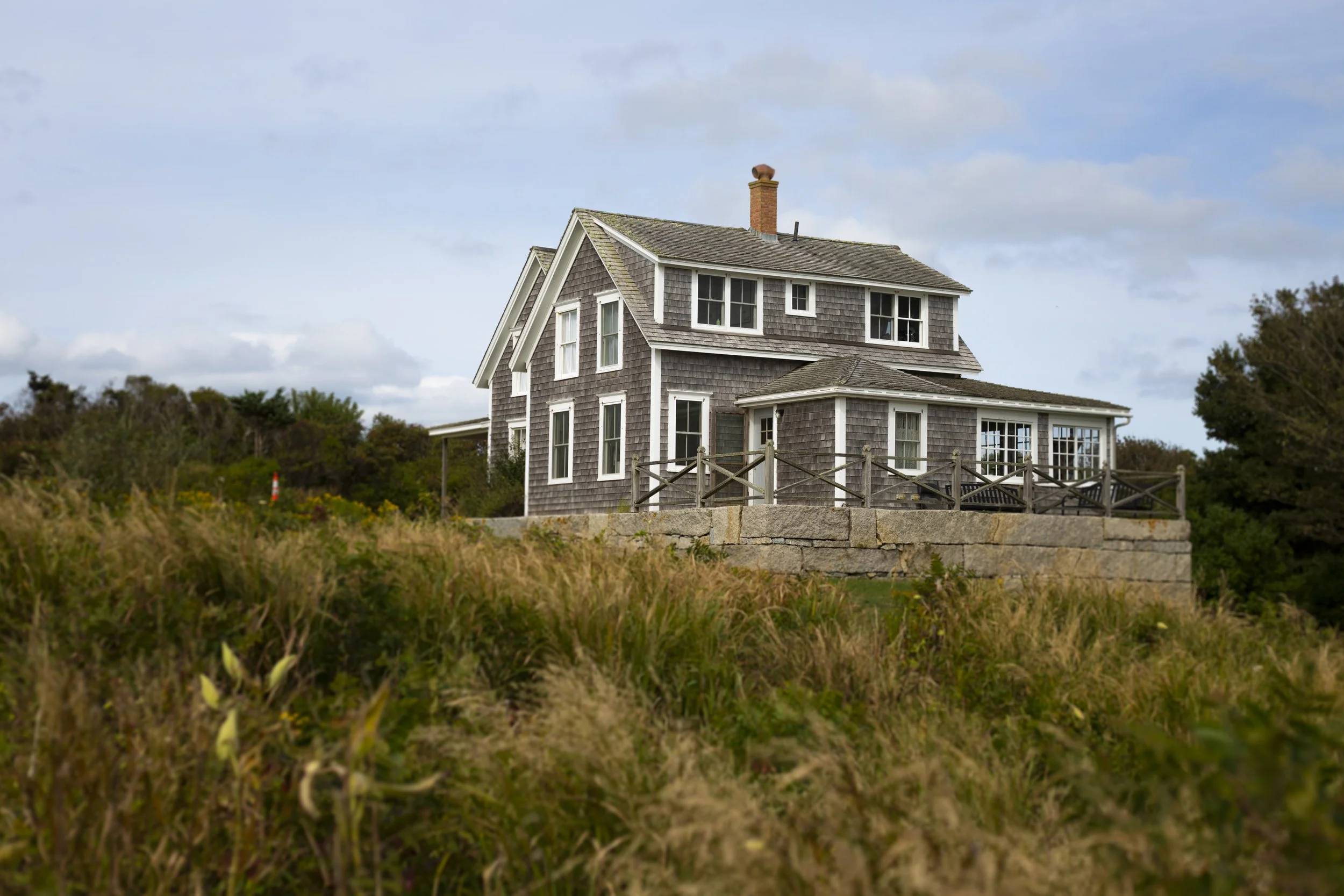 A multi-story house with gray shingle exterior, white trim, multiple windows, a kitchen chimney, and a small deck surrounded by a wooden fence, set against a partly cloudy sky and greenery.