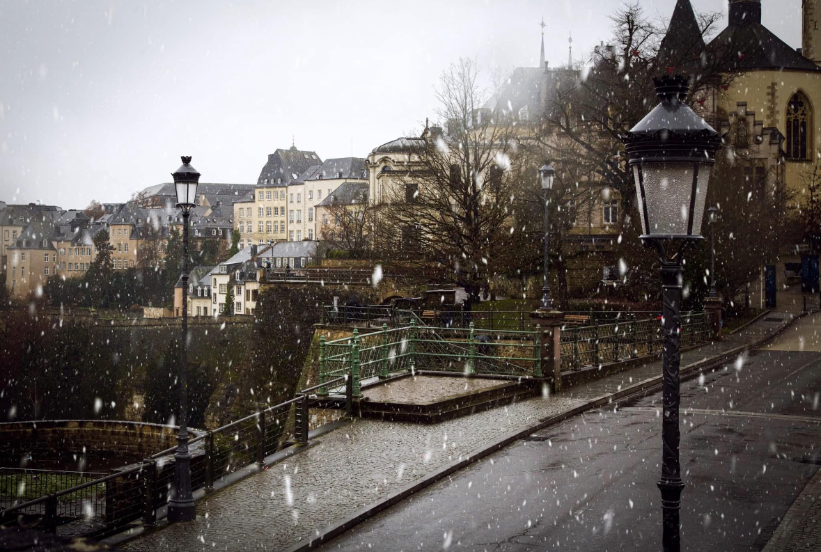 A snowy street scene with old European-style buildings, street lamps, and trees.