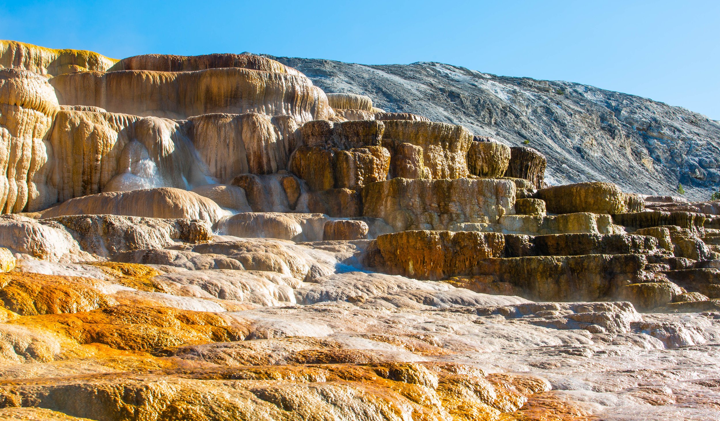 Hot springs with mineral deposits creating colorful terraces on a mountainside under a clear blue sky.