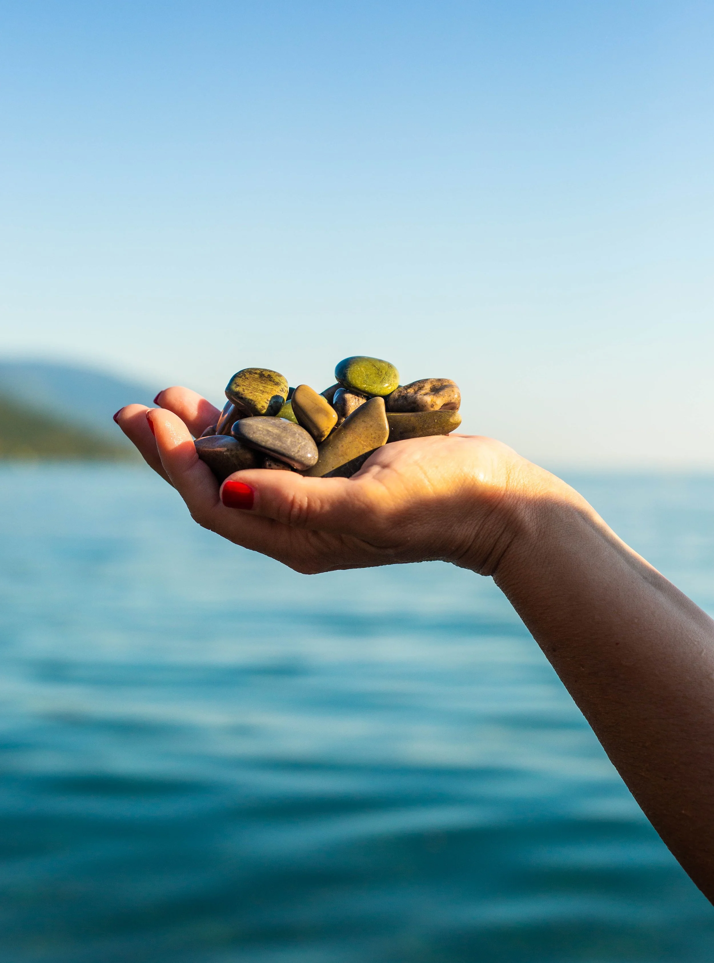 A hand holding a small pile of smooth, colorful rocks against a blurred body of water and sky background.