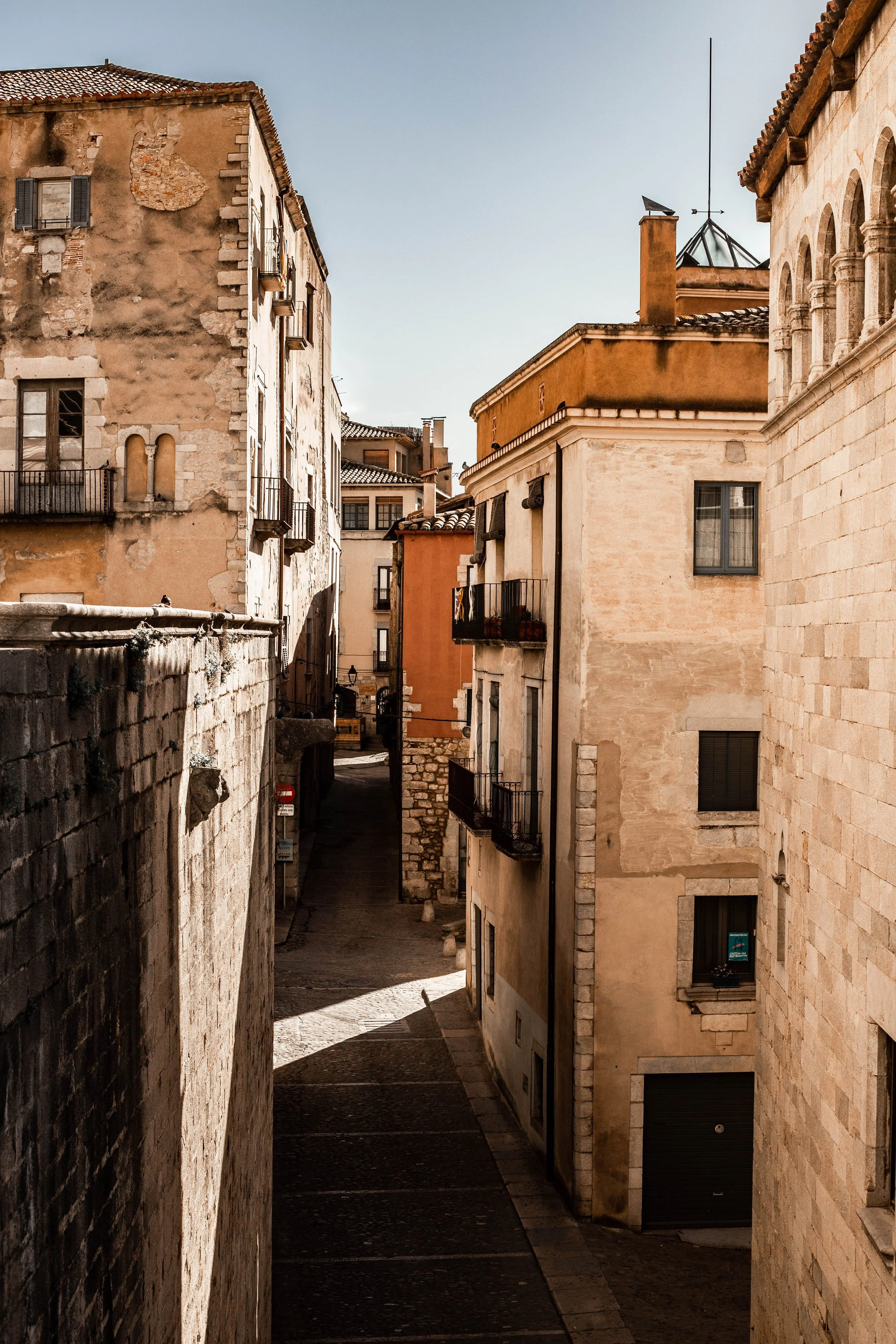 Narrow street lined with old, weathered buildings with balconies, windows, and stone walls, in warm sunlight.