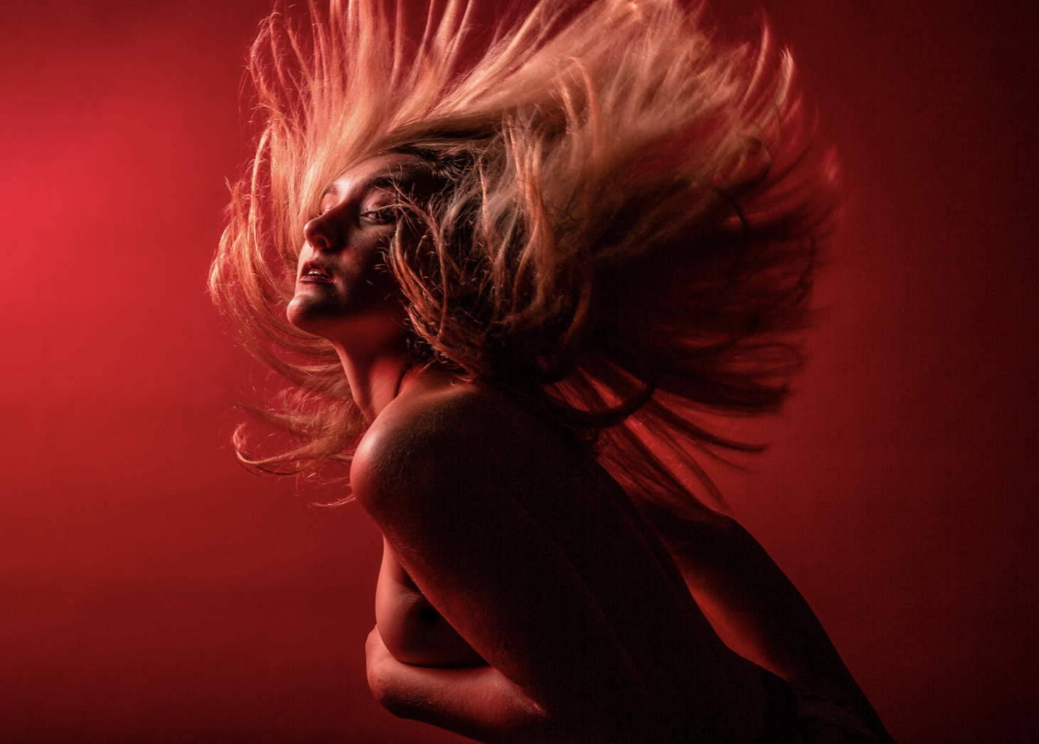 A woman with long flowing hair captured in a dramatic pose against a red background, with intense lighting highlighting her face and shoulders.