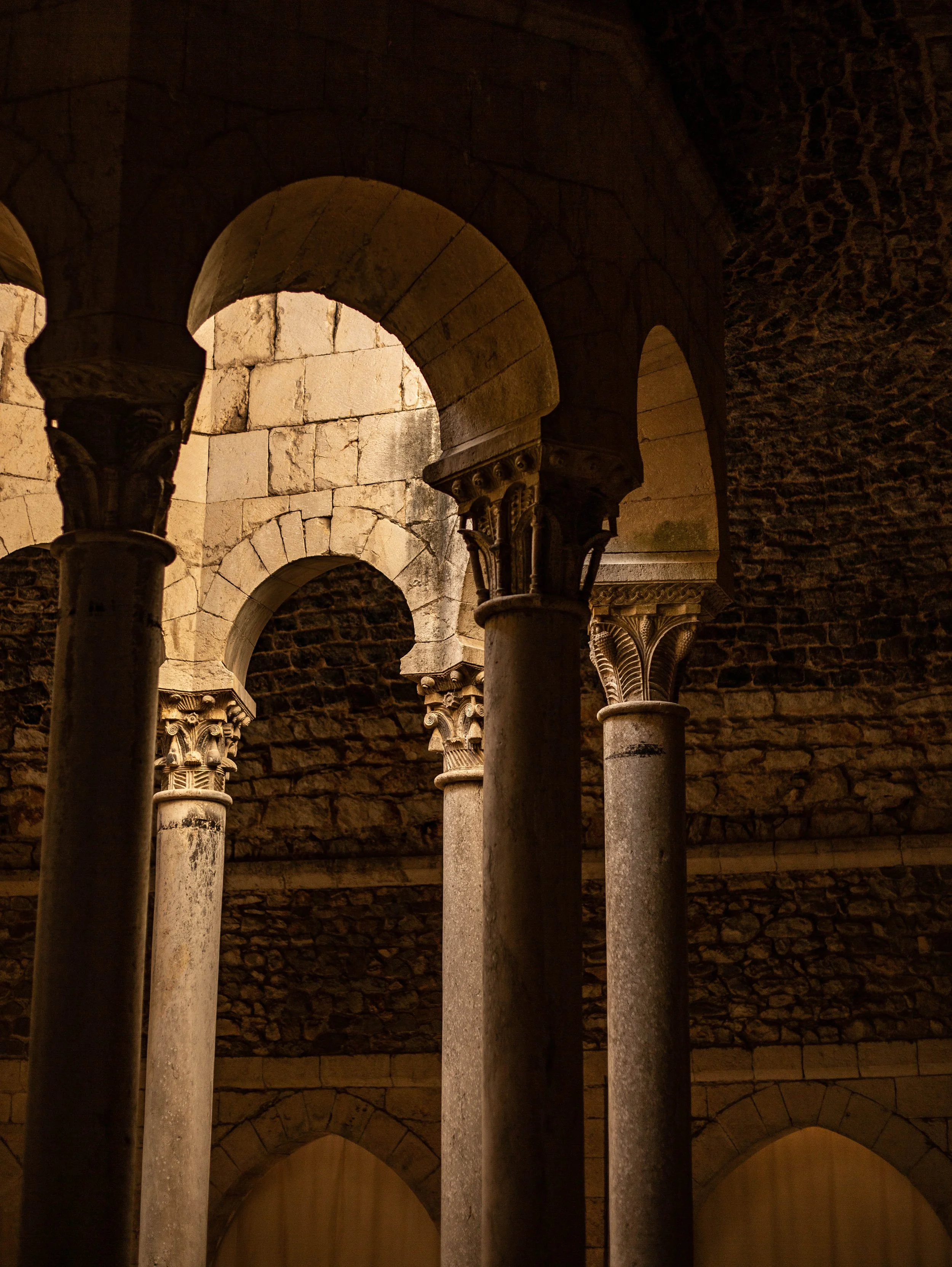 Ancient stone archways and columns inside a historical building, with warm lighting highlighting the textured stone walls and architectural details.