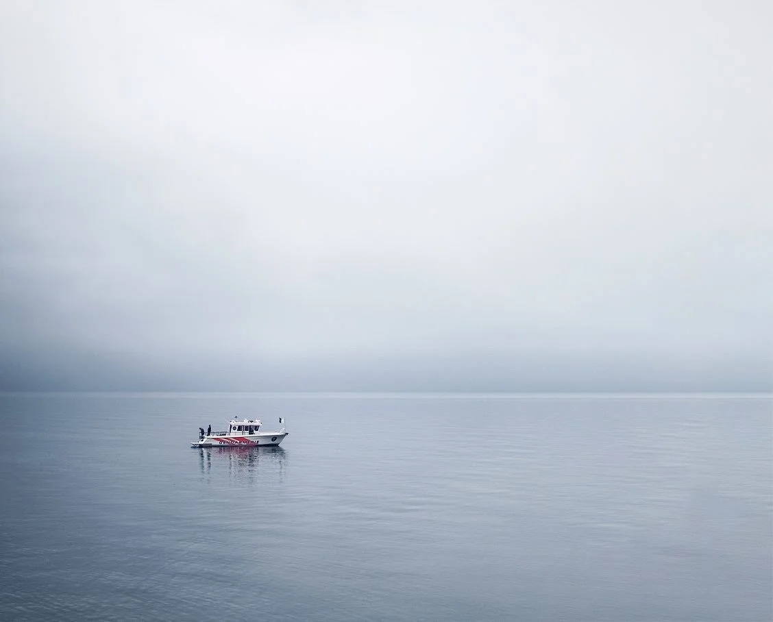 A small boat on a calm, expansive body of water with a foggy, gray sky in the background.
