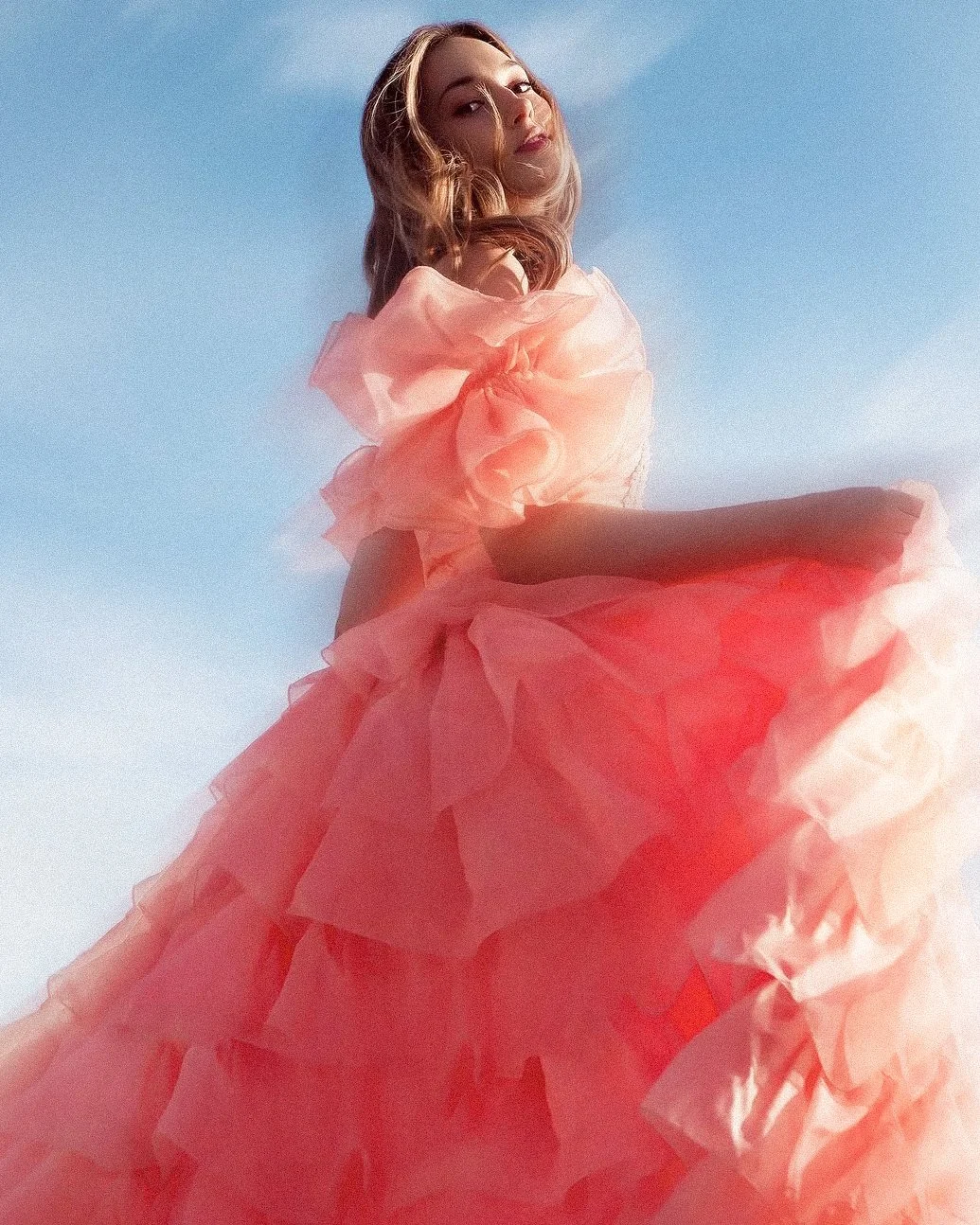 A woman in a voluminous pink ruffled gown standing outdoors against a clear blue sky.