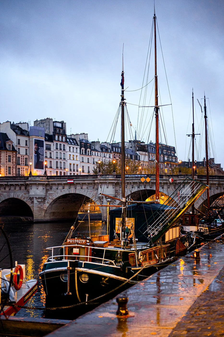 A docked boat on a river with a city bridge and multi-story buildings in the background, during overcast weather.