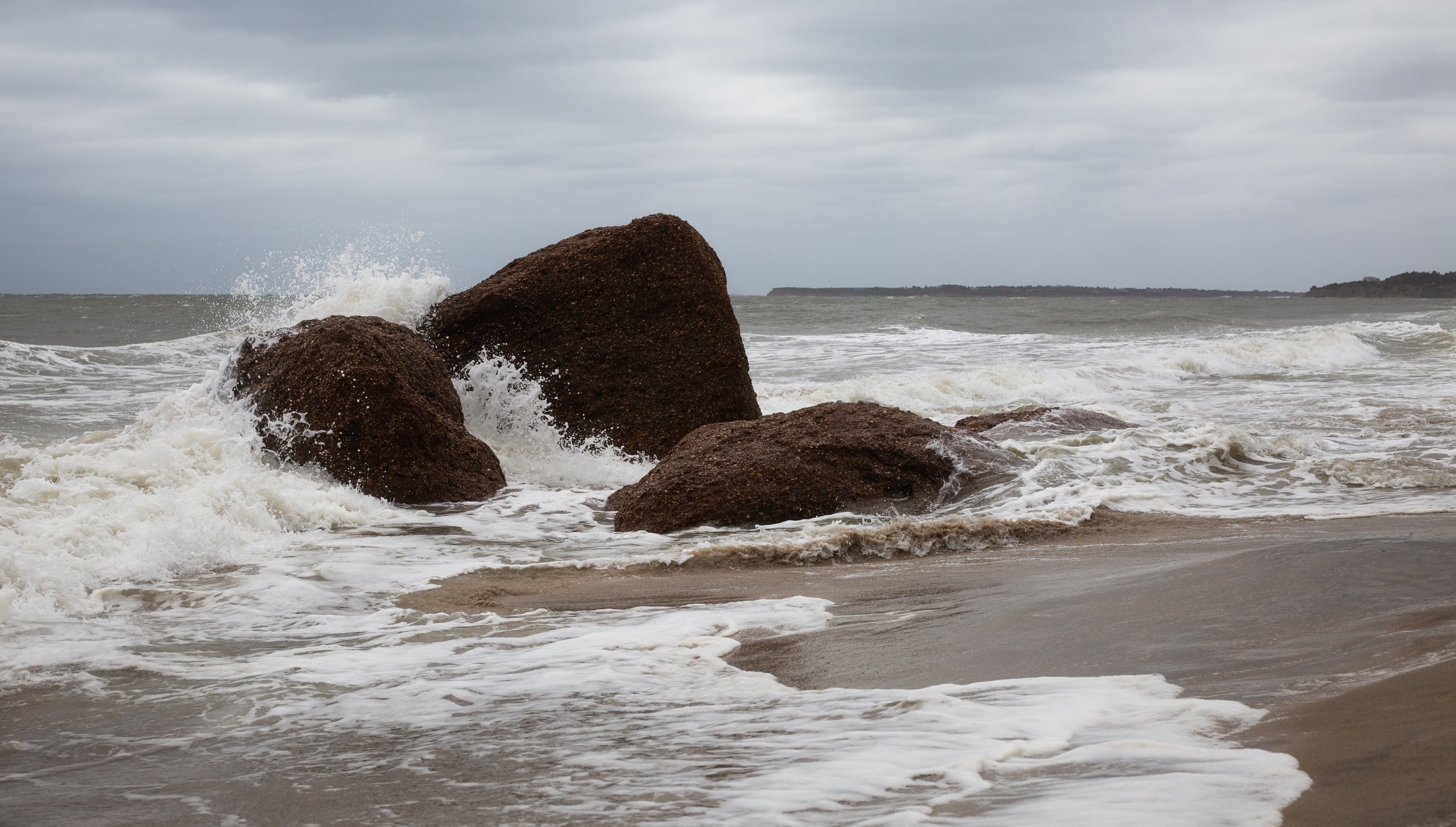 Large rocks in the ocean with waves crashing against them on a cloudy day.