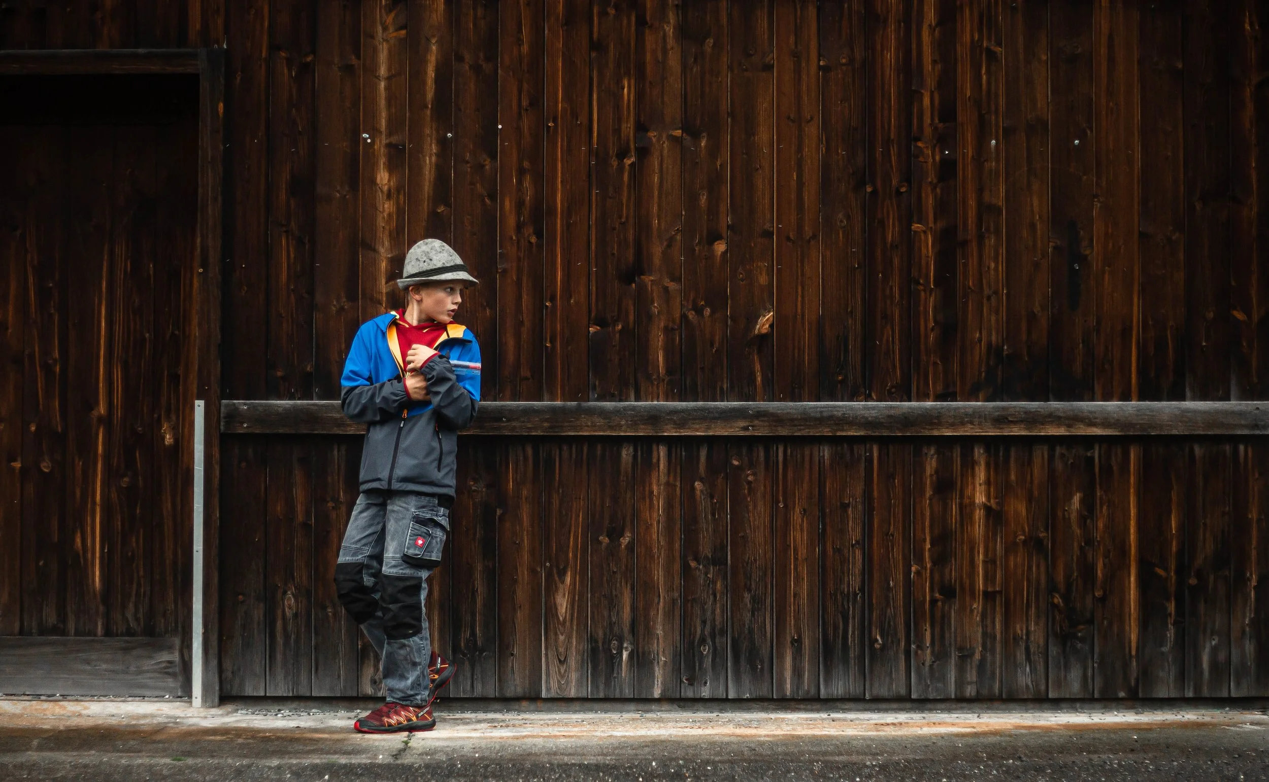 Young boy in colorful outdoor jacket and gray hat leaning against a wooden fence.