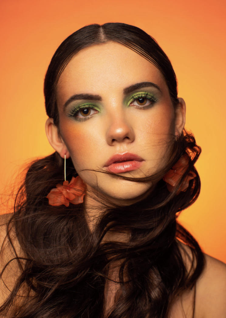 Close-up portrait of a young woman with long dark hair, green eyeshadow, orange flower earrings, and a warm orange background.