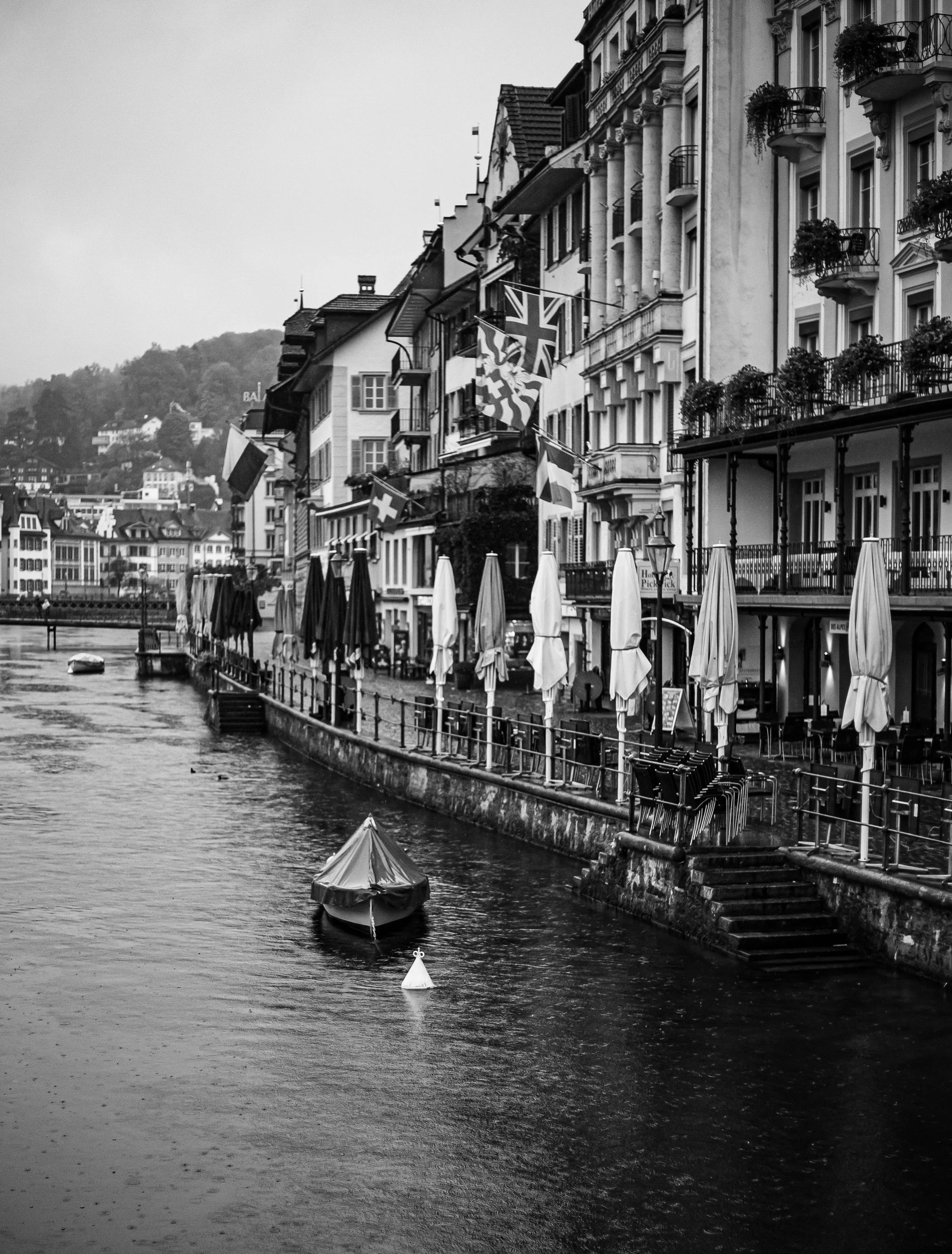 Black and white photo of a European city waterfront with buildings displaying flags from various countries, including the United Kingdom and Switzerland. A small boat is floating in the water, and there are umbrellas and chairs along the waterfront. 