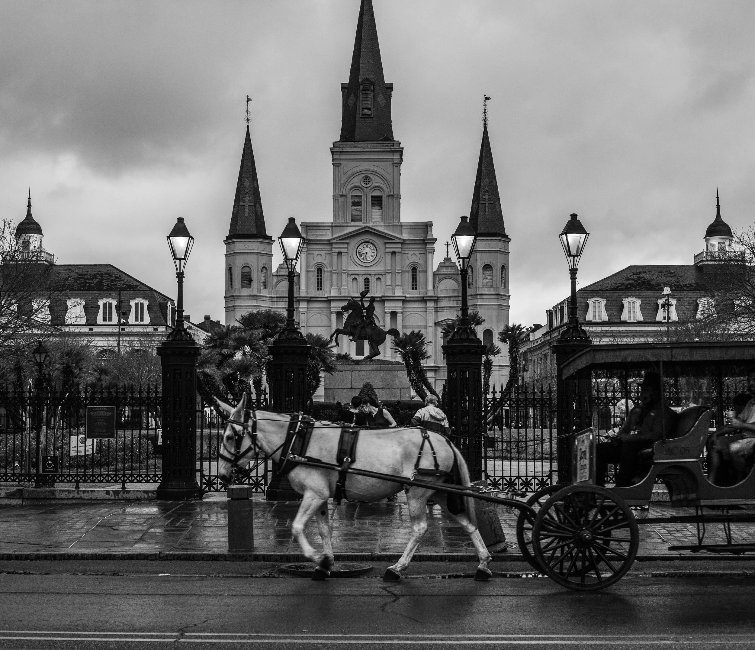 A black and white photograph of a church with multiple spires, a clock on its facade, and a statue in front. There are street lamps and horse-drawn carriages passing by, with overcast skies above.