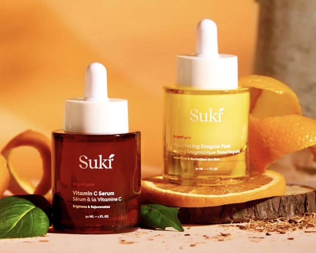 Two bottles of Suki skincare products, Vitamin C Serum in a red bottle and BrightC Eye & Face Enzyme Peel in a yellow bottle, surrounded by orange slices and green leaves.