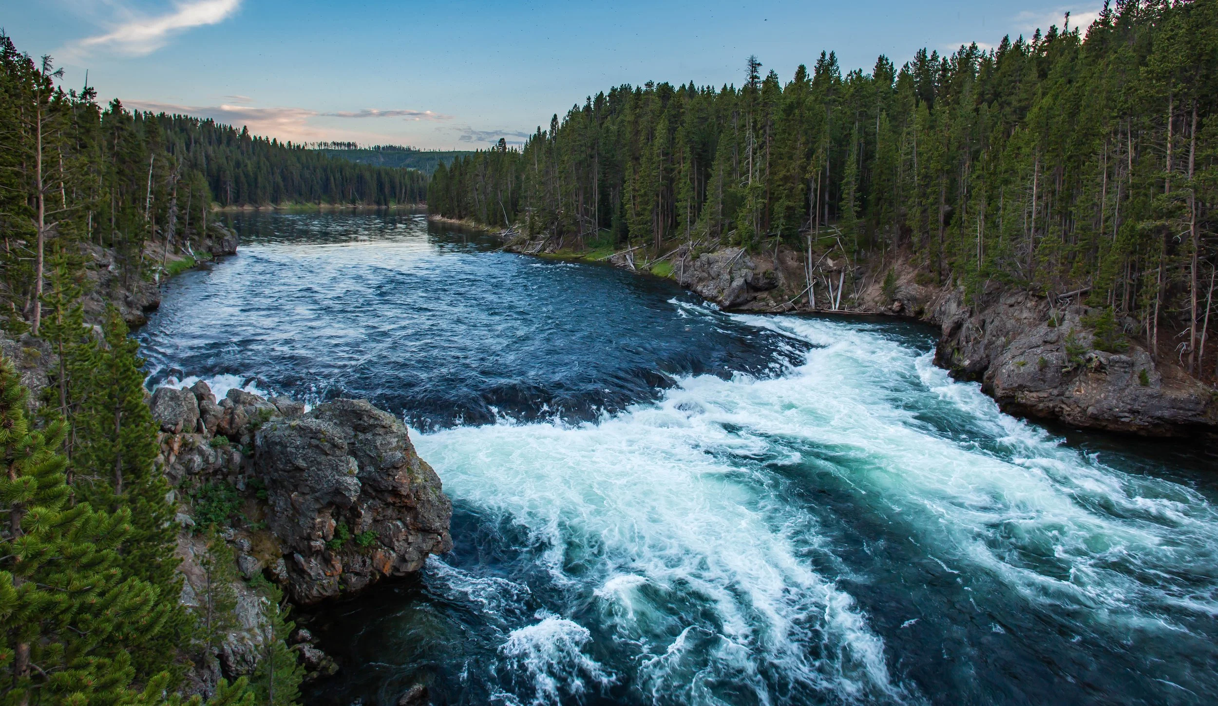 A river flowing through a forested canyon with tall evergreen trees and rocky shores at sunset.