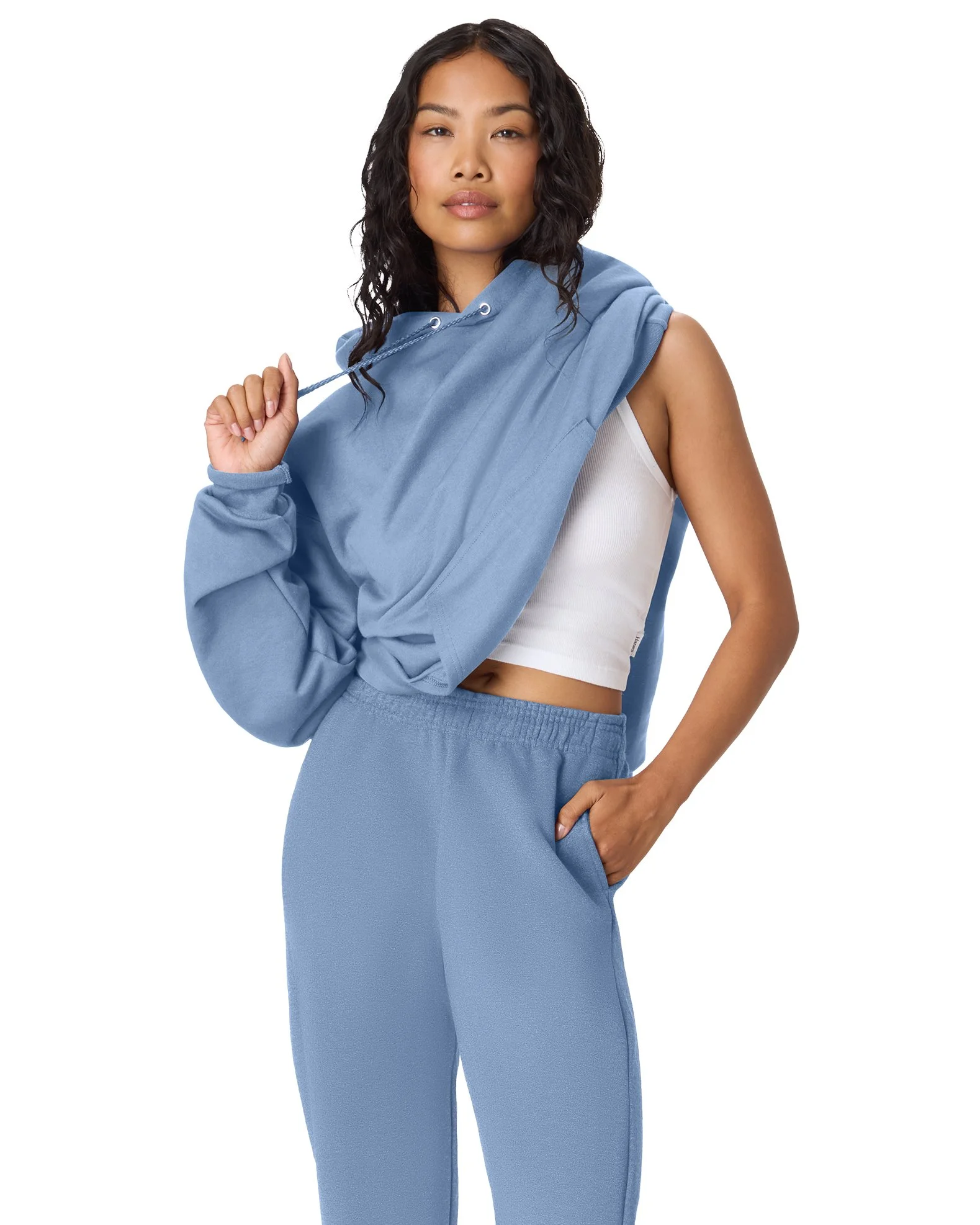 Woman with wavy black hair wearing a blue hoodie and matching blue sweatpants, standing with one hand in her pocket and the other holding the hoodie drawstring, against a plain white background.