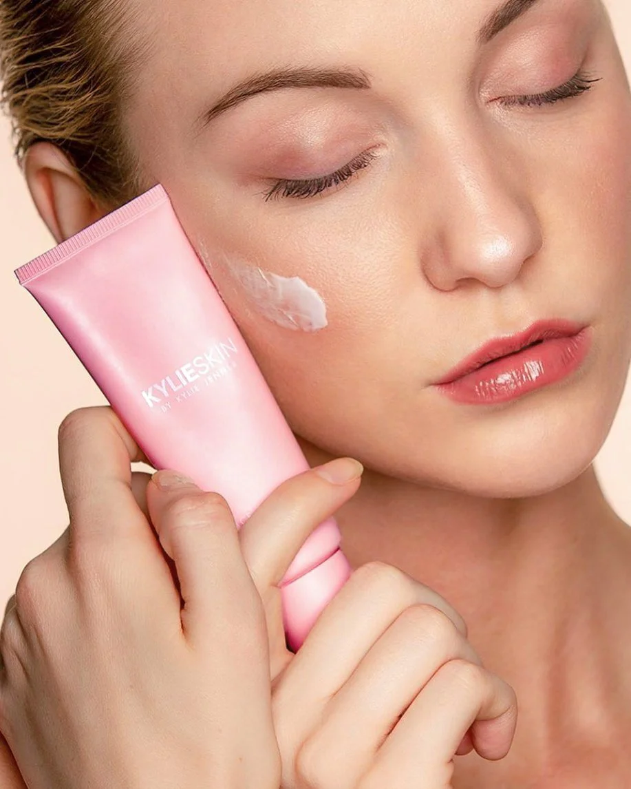 A woman with closed eyes applying a cream or moisturizer from a pink tube labeled 'KYLIESKIN' to her cheek.