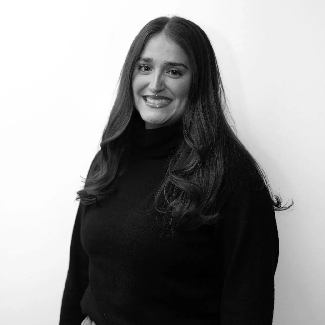 Black and white photo of a woman with long hair, smiling and wearing a black top and a thin necklace, against a plain dark background.