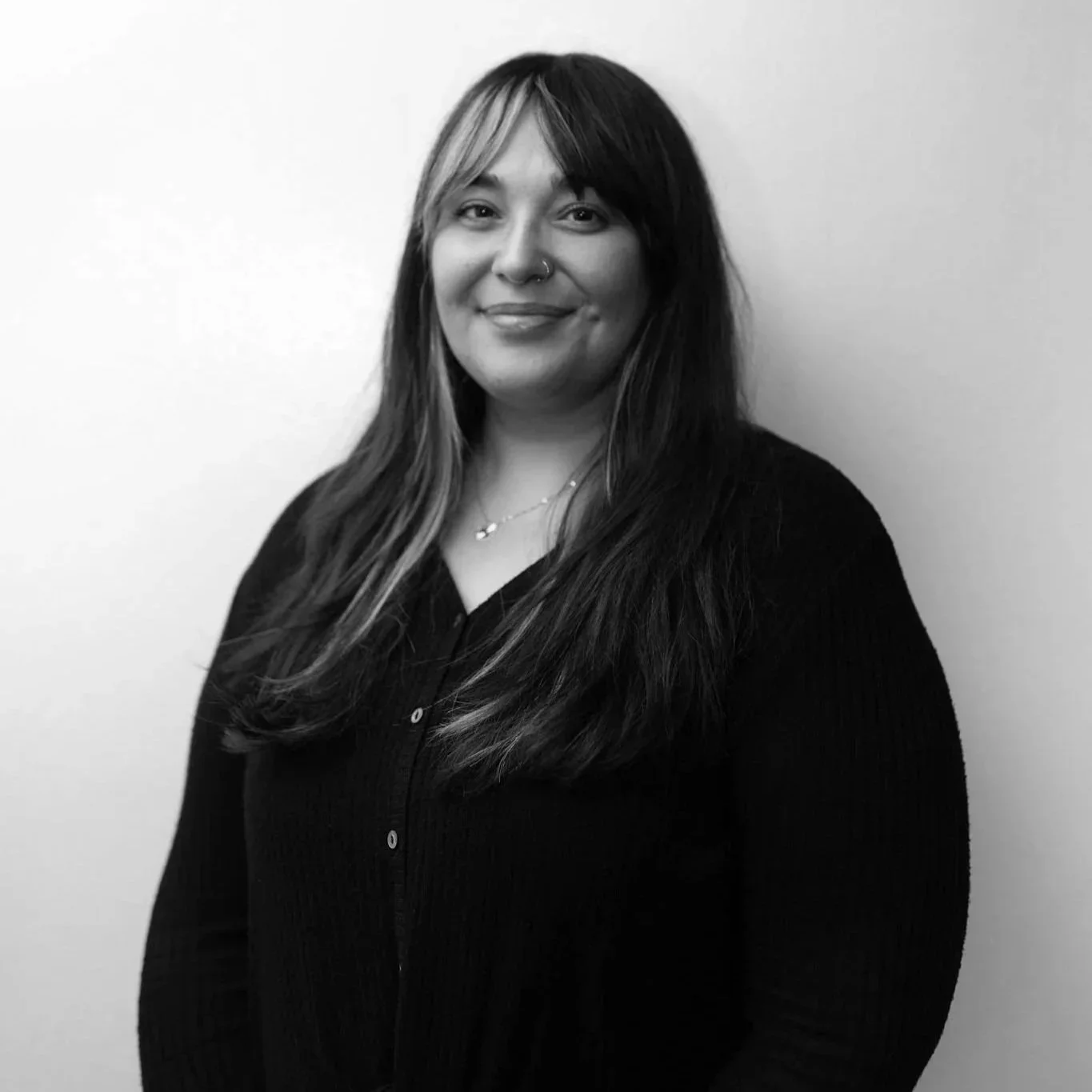 Black and white photo of a smiling woman with glasses, wearing a black V-neck shirt and a necklace, standing against a dark textured background.